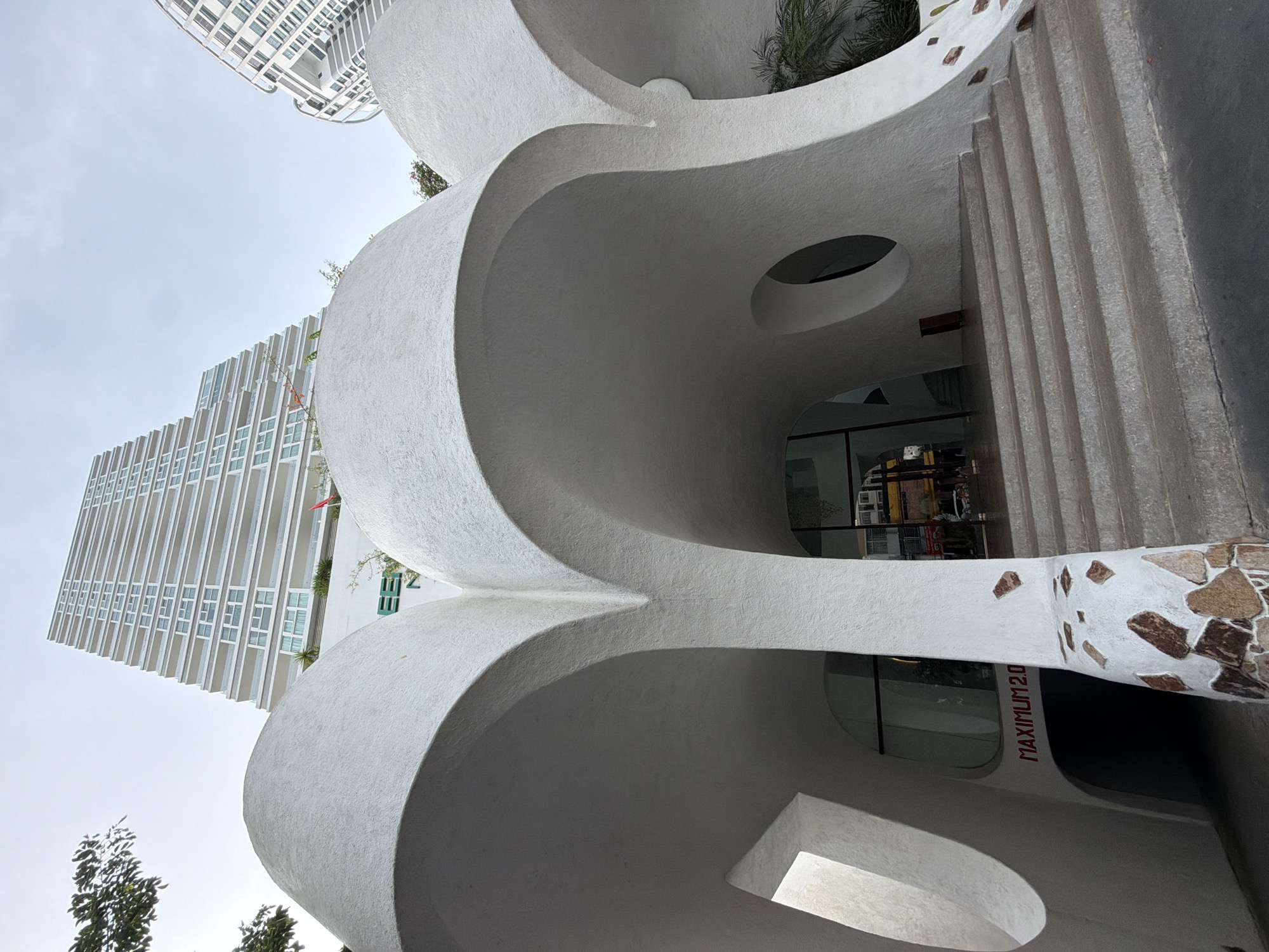 The image displays an urban architectural scene, likely in Nha Trang Ward, Vietnam, under a bright, clear daytime sky. In the foreground, a unique, multi-layered white building dominates the lower and central parts of the frame. It features an organic, sculptural design with flowing, undulating curves and a textured, possibly stuccoed, surface. This structure incorporates various openings and cut-outs, including a circular one, and creates dynamic interplay of light and shadow on its complex forms. Towards the bottom right, a section of this building's exterior is adorned with irregular, earthy-toned stone cladding. Adjacent to this, textured steps or a ribbed wall ascend on the right.
In the mid-ground and background on the left, a tall, modern high-rise building stands, presenting a stark contrast with its rigid, rectangular form, multiple windows, and balconies arranged in a grid pattern. Patches of green foliage and trees are visible in various parts of the frame, hinting at landscaping around the buildings. No people are visible. The bright, even lighting suggests clear weather, possibly morning or afternoon. On the white, curvilinear building, near an entrance or window, the red text "MAXIMUM 2.0" is clearly visible.