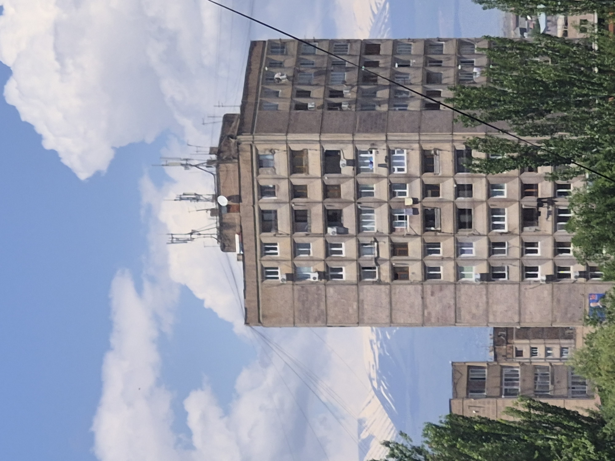 The image displays a towering, multi-story building, likely an apartment block, featuring a grey concrete brutalist architectural style. The building's facade is characterized by a grid pattern of recessed square windows and small balconies, some of which are open or have white frames. Several air conditioning units are visible beneath some windows. The flat roof of the building is adorned with multiple communication antennas and at least one satellite dish, suggesting a dense urban environment.
The structure is set against a vibrant blue sky, dotted with large, fluffy white clouds. In the background, beyond the main building, faint outlines of distant mountains or hills can be observed beneath the cloud line. Green, leafy trees partially obscure the lower right and bottom left portions of the building, indicating a treed street or park area nearby. Black utility wires diagonally traverse the upper part of the image.
The scene is captured in bright daylight, suggesting mid-morning or afternoon, with clear weather. No specific human activity is visible, and the overall impression is one of a static urban landscape. There is no discernible text visible in the image. The architectural style strongly suggests a location within a former Soviet bloc country, aligning with the provided context of Yerevan, Armenia.