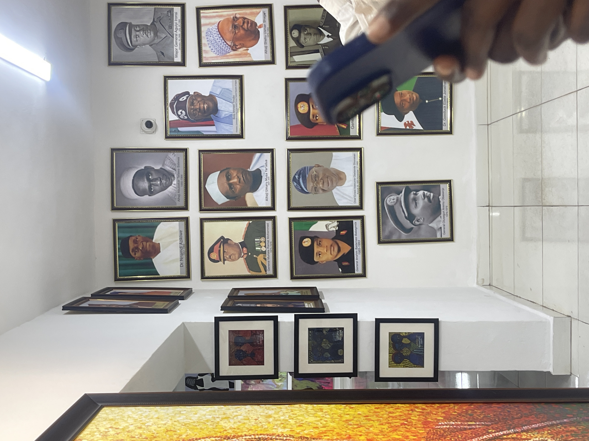 This media file depicts an indoor setting, likely a gallery, museum, or an institutional office in Abuja, Nigeria. The white walls are adorned with numerous framed portraits of prominent Nigerian leaders and historical figures, arranged in multiple rows. The floor is covered with light-colored tiles.
In the upper right corner, the hand of a person holding a smartphone is visible, suggesting an activity of taking a photograph or scanning one of the exhibits.
A long fluorescent light fixture is mounted on the upper left wall, illuminating the space, and a small, white security camera is installed on the wall below it.
The framed portraits showcase a mix of black and white and color images. Visible text on the portraits identifies figures such as:
- Major General Aguiyi Ironsi (January 15, 1966 - July 29, 1966)
- Alhaji Abubakar Tafawa Balewa (1960-1968)
- General Murtala Ramat Mohammed (1975-1976)
- Alhaji Umaru Musa Yaradua
- General Ibrahim Badamasi Babangida (1985-1993)
- Chief Ernest Adekunle Oladeinde Shonekan (August 26, 1993)
- General Olusegun Aremu Obasanjo (1976-1979)
- Bola Ahmed Tinubu
- Dr. Goodluck Ebele Jonathan
- Alhaji Shehu Usman Shagari (1979-1953)
- Dr. Nnamdi Azikiwe (1963-1966)
Below the main display of portraits, on a white ledge or shelf, several smaller framed artworks featuring abstract patterns and designs are showcased. At the bottom of the image, a large, vibrant artwork or painting with warm hues of yellow, orange, and red is partially visible. The overall scene suggests a historical or commemorative exhibition space.