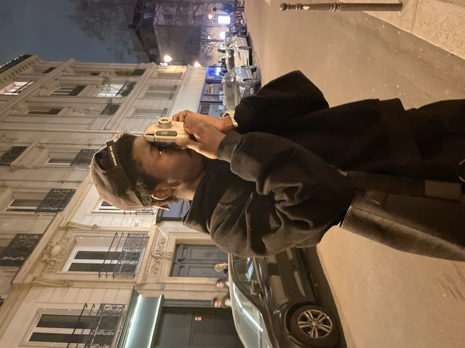 An individual with dark skin, wearing a black hoodie and a dark baseball cap with "PROJECT GR" text, is depicted in a medium shot on a street in Paris, France, at night. The person holds a light-colored, possibly beige or silver, point-and-shoot camera to their right eye, appearing to frame a photograph. A dark strap from a bag crosses their torso. The background features classic Haussmannian multi-story stone buildings with ornate wrought-iron balconies, characteristic of Parisian architecture. A dark sedan is partially visible parked on the left, with other vehicles lining the street further in the distance. Blurred figures of additional individuals are discernible in the left background. The scene is illuminated by artificial light sources, including streetlights and building lights. A dark bollard stands on the right foreground.