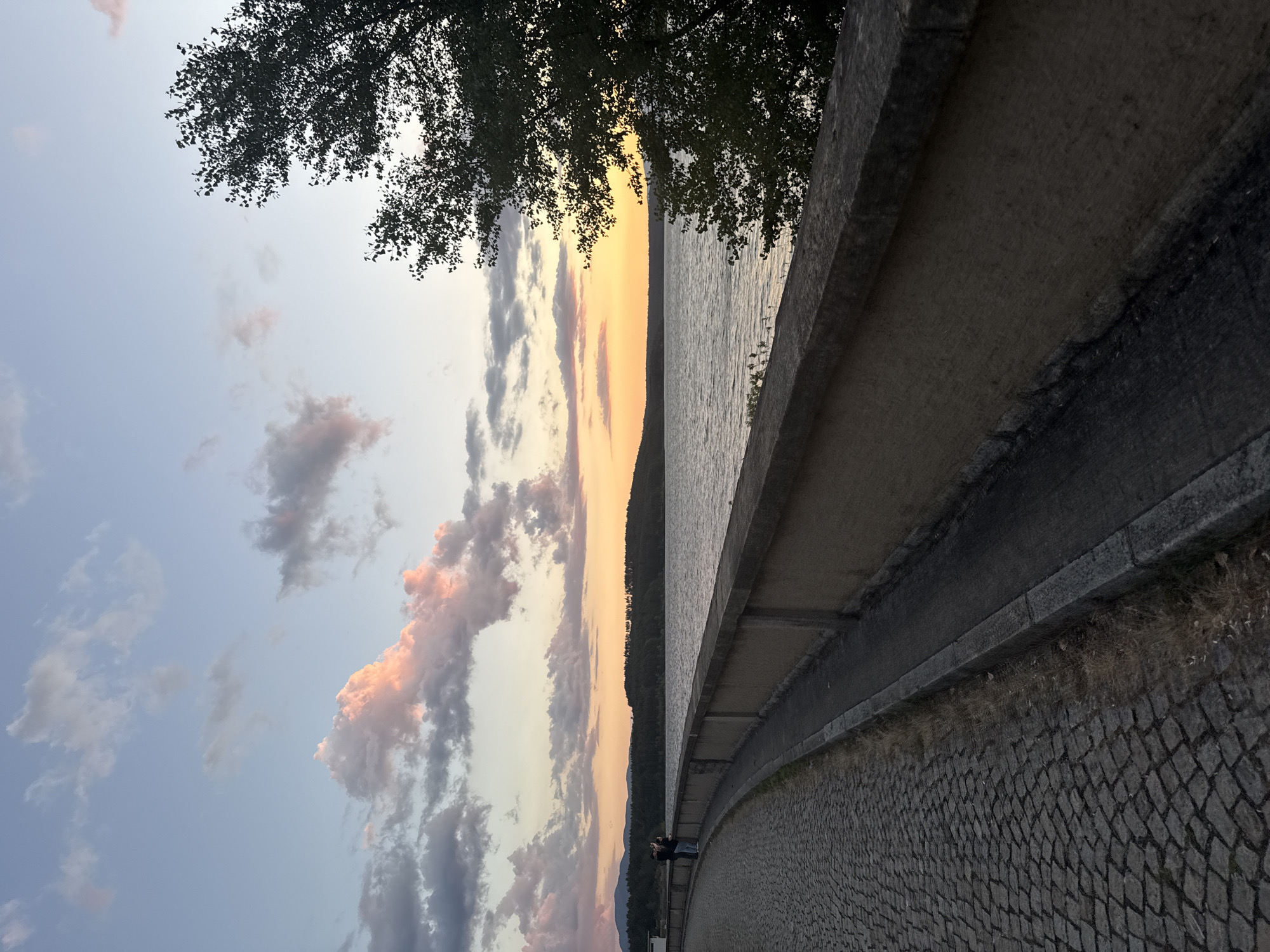 The image depicts a scenic view of the Koprinka dam in Koprinka, Bulgaria, at sunset. A cobblestone pathway curves along the edge of the dam, bordered by a stone wall. On the pathway, two individuals stand facing the expansive lake, seemingly admiring the sunset. The water of the lake reflects the warm hues of the sky, which is adorned with scattered clouds tinged with pink and orange. The background features a treeline and distant hills, silhouetted against the colorful sky. A large tree with dark foliage is visible on the right side of the frame, framing the view. The overall atmosphere is tranquil and picturesque, capturing the beauty of nature at dusk.