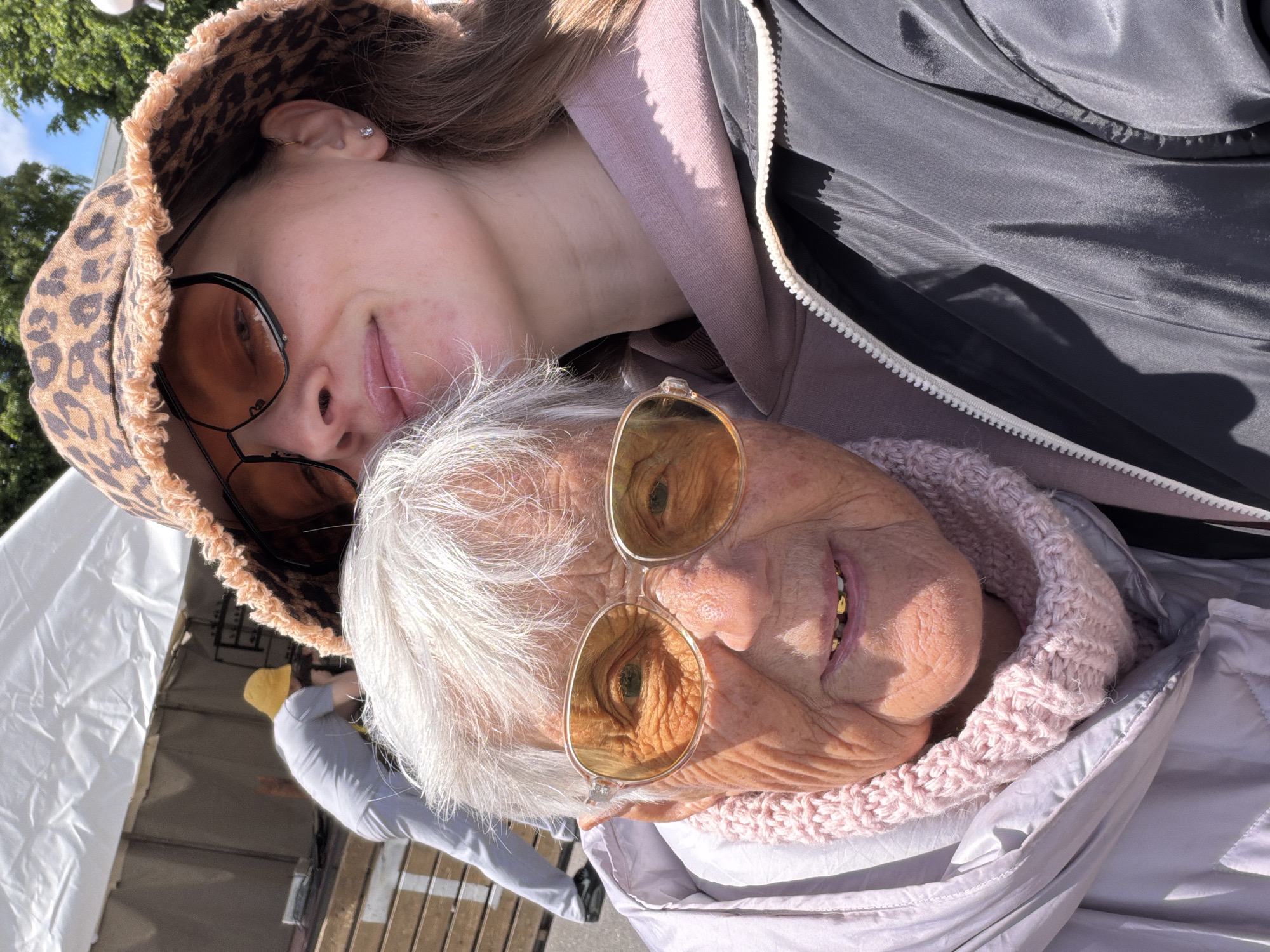The image shows two people posing closely together. The younger person, wearing a patterned bucket hat and tinted glasses, is positioned slightly behind the older person. The older person, also wearing tinted glasses, has white hair and a pink knitted scarf. A gray zippered jacket is visible. The background includes a white tent-like structure and a glimpse of a person in a gray shirt and yellow hat. The location, Valozhyn, Belarus, is not verifiable based on the image content alone.