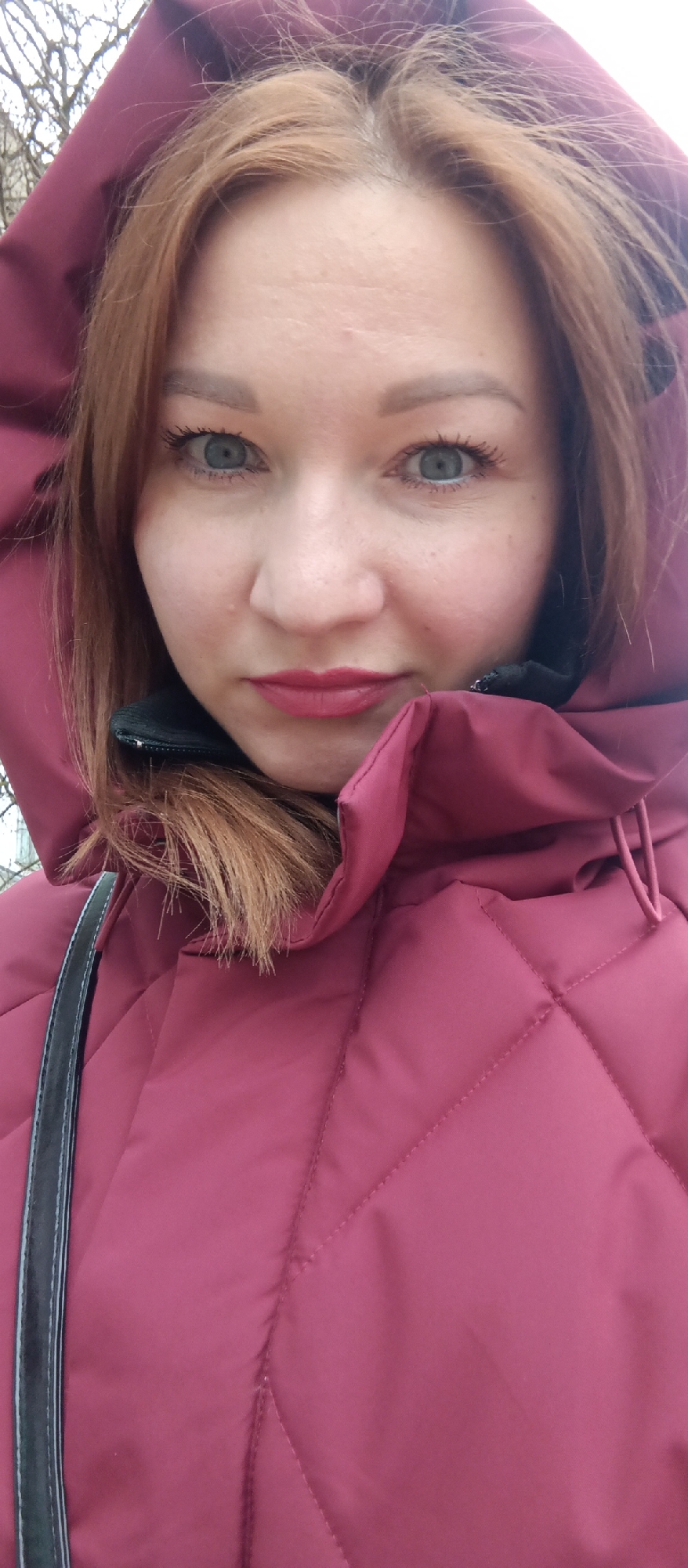 A close-up shot depicts a woman with fair skin, reddish-brown hair, and light blue eyes. She is wearing a quilted, deep red jacket with the hood pulled up, framing her face. A black strap of a bag or purse is visible over her left shoulder. The background shows some blurry foliage, suggesting an outdoor environment. The photo appears to be taken during the daytime with natural lighting. Her expression is subtle, with a slight smile. The location is an urban environment, possibly a park or street in the city of Unknown, Unknown.