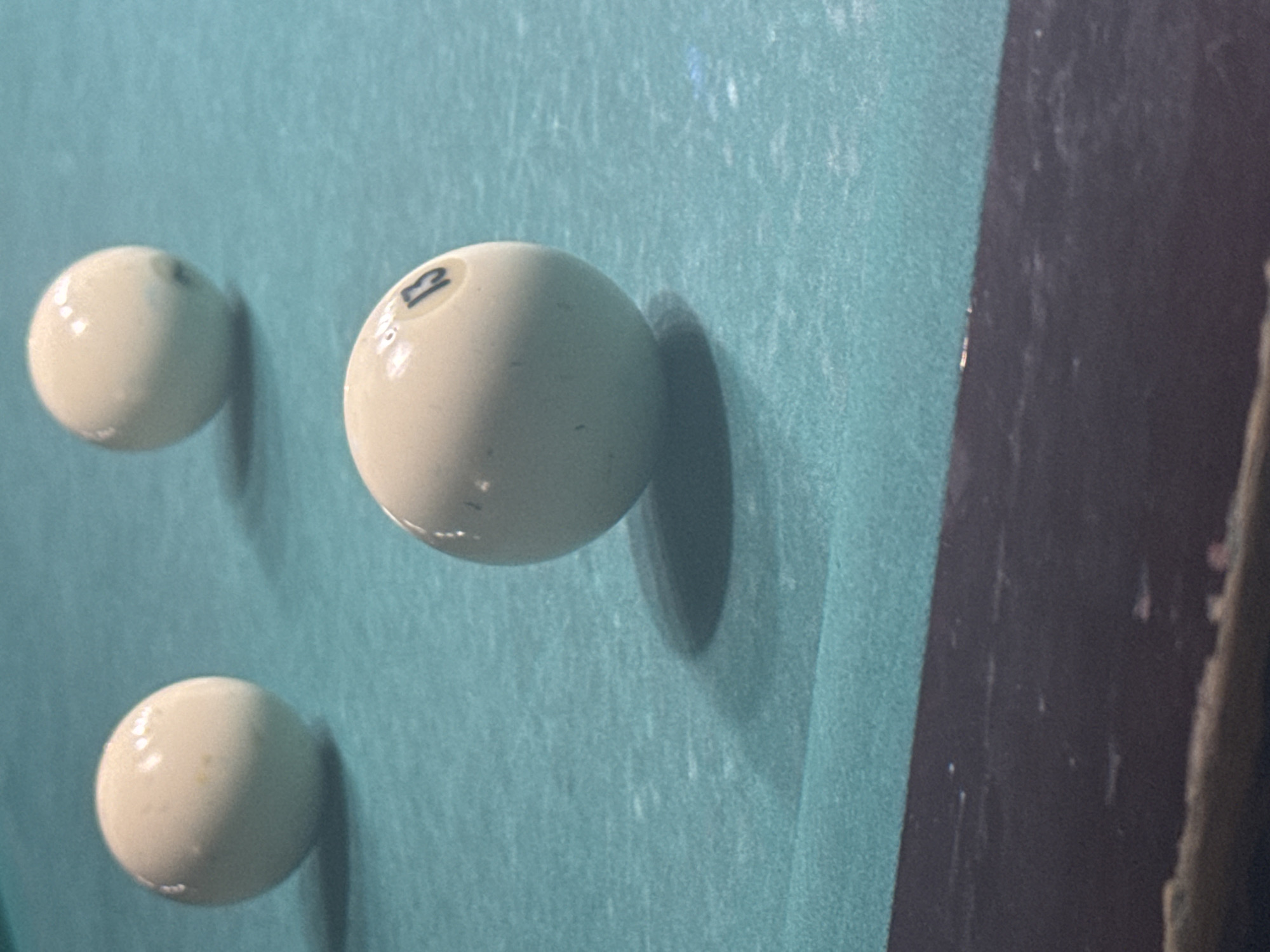 The image shows three off-white billiard balls on a turquoise billiard table. The center ball has the number "13" on it. A portion of the table's dark-colored side rail is visible on the right. No people are present. The location is not identifiable from the image.