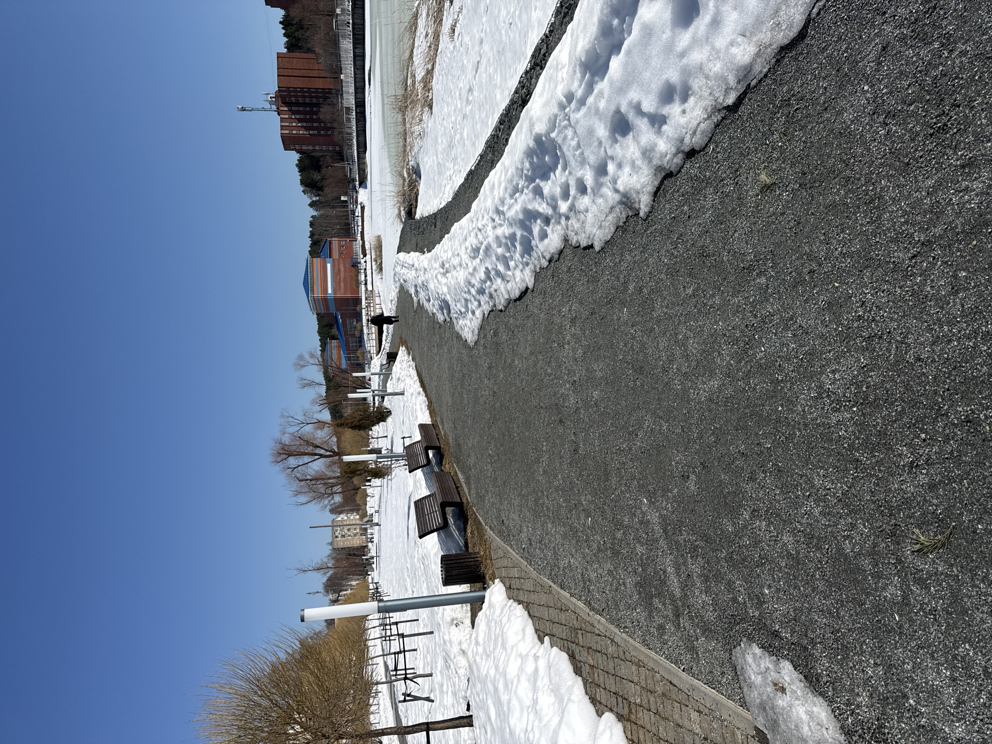 The image depicts a path in Votkinsk, Russia, partially covered with snow, suggesting a winter or early spring scene. The path is paved with small, dark gravel and is flanked by snow on both sides. In the background, buildings are visible, including what appears to be a high-rise apartment building and a structure with a blue roof. Benches and lamp posts line one side of the path. A solitary figure can be seen walking in the distance. The sky is clear and blue, indicating a sunny day.