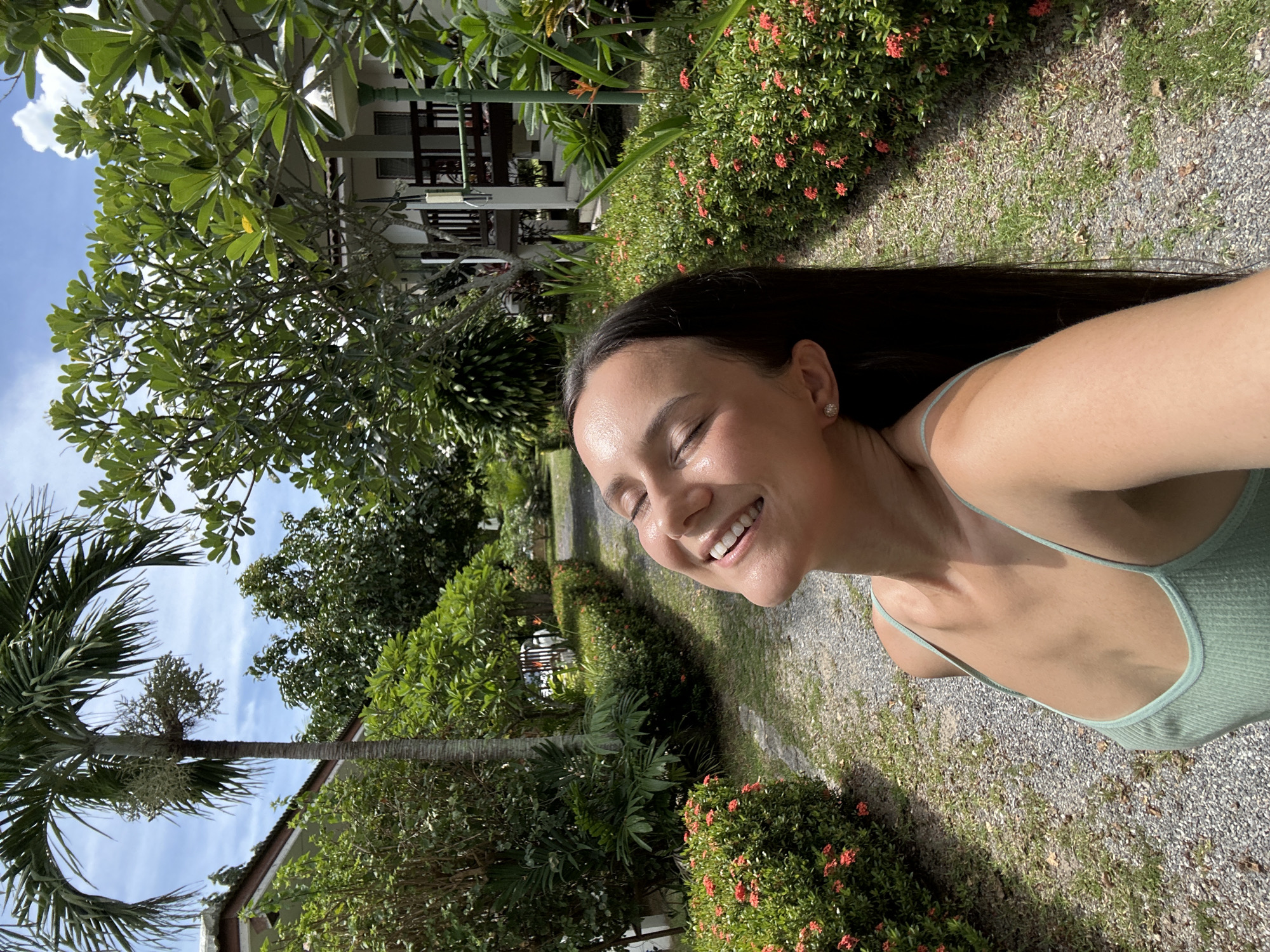 A woman with dark hair and fair skin is pictured in a selfie taken outdoors in Baan Mae Nam, Thailand. She is wearing a light green spaghetti strap tank top and a diamond stud earring. Her eyes are closed, and she is smiling with her head tilted back, basking in sunlight. She appears to be standing on a gravel path lined with lush green bushes and vibrant red ixora flowers. Palm trees and tropical foliage are visible in the background, along with a building with a white exterior and a dark-pitched roof. The sky is bright blue with scattered white clouds.