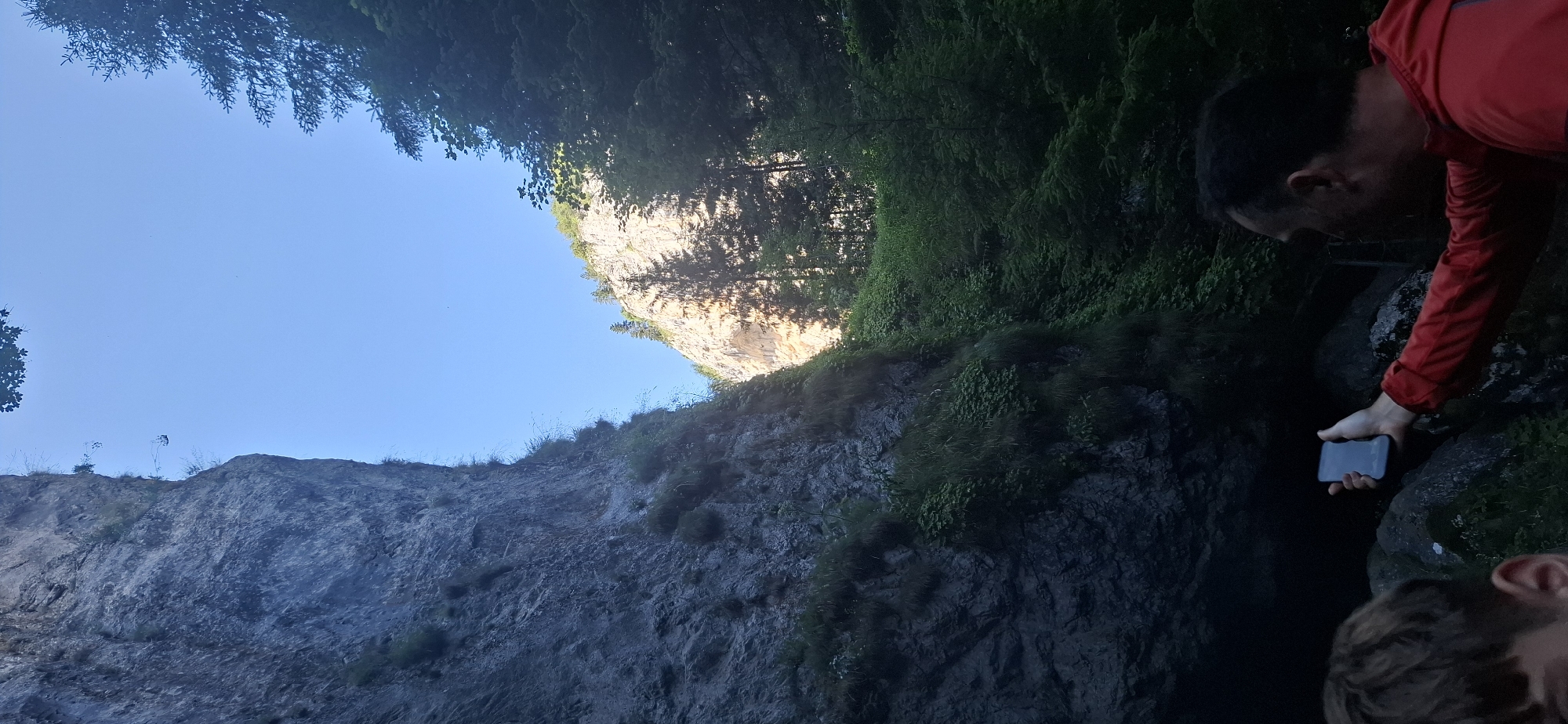 The image depicts a narrow canyon with steep, rocky walls on either side. Dense green foliage covers parts of the lower canyon walls and extends up the right side of the canyon, where tall evergreen trees are visible. The sky is a clear, bright blue, with sunlight illuminating the upper portion of the right canyon wall and filtering through the trees. In the foreground, a person is visible from the chest up, wearing a red jacket and holding a smartphone in their right hand, appearing to take a picture or video. Part of another person's head and shoulder, with light brown hair, is visible in the lower left corner of the frame. The scene is consistent with the Trigrad Gorge in Trigrad, Bulgaria, known for its dramatic geological formations.