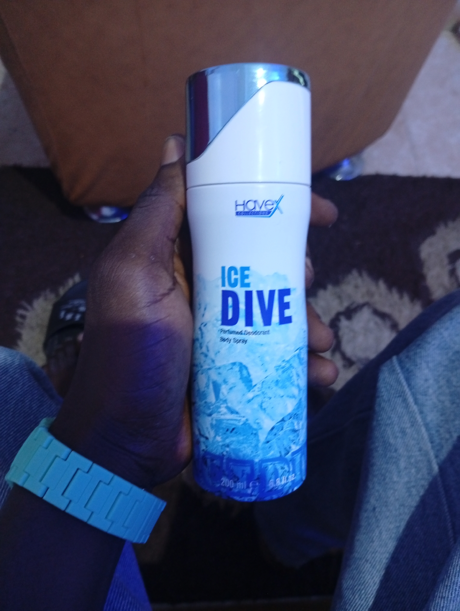 A dark-skinned hand holds a white and silver Hovex Ice Dive deodorant body spray. The person is wearing a blue shirt and a blue watch. The background includes a brown surface and a patterned rug. The location is unknown, the city of Lokoja, Nigeria, is not evident.