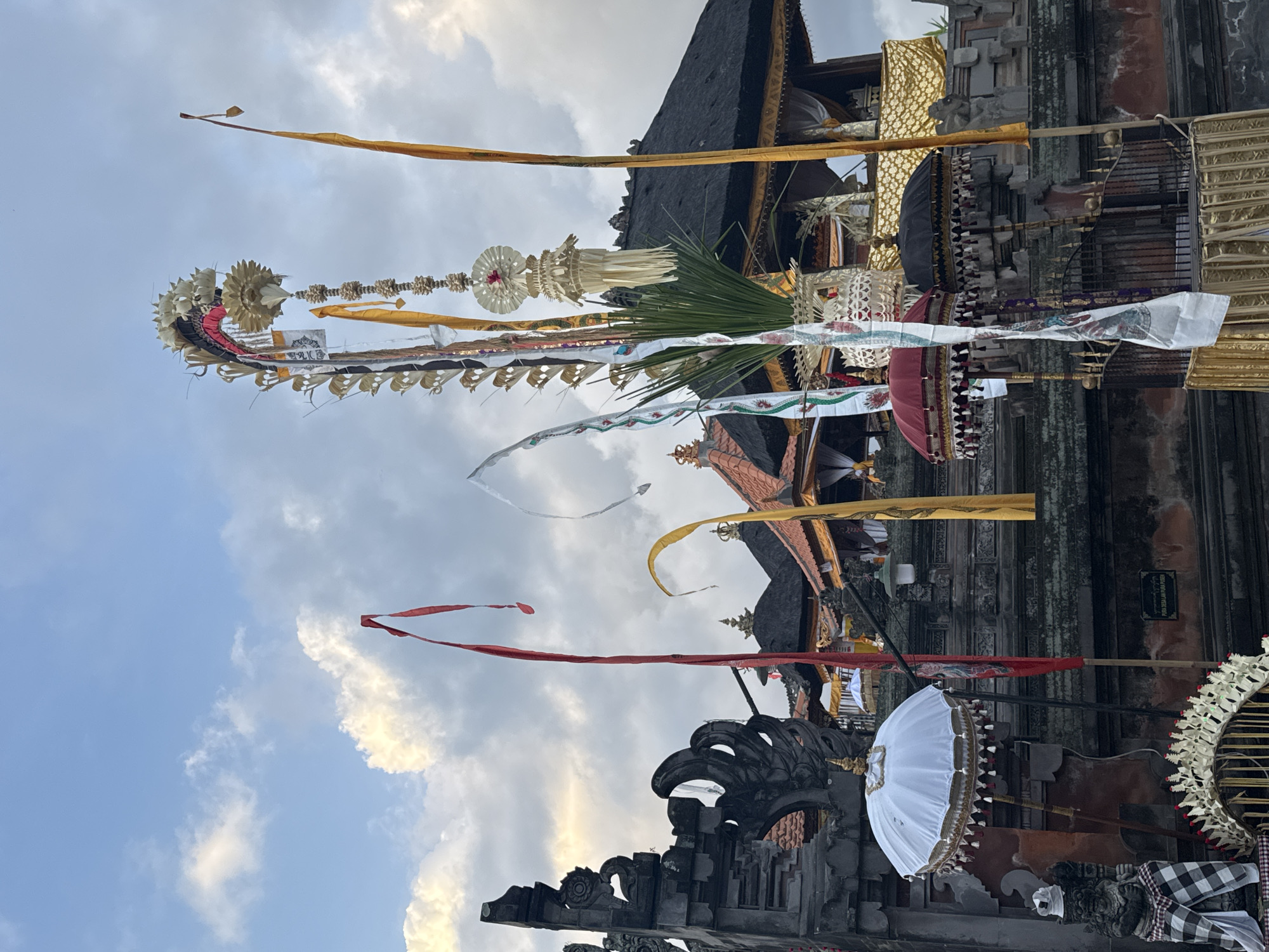 A wide shot captures a street view in Blahbatuh, Indonesia, with ornate Balinese temple architecture dominating the scene. Multiple tall, decorative banners, some adorned with intricate carvings and colorful fabrics, stand prominently. Red, yellow, and white flags flutter gently in the breeze. At the lower left, a traditional Balinese stone statue is draped in a black and white checkered cloth. A white ceremonial umbrella with gold trim stands to the right of the statue. In the background, a temple structure with a dark, thatched roof and red accents is visible, flanked by other buildings featuring intricate stonework and decorative elements. Several smaller, covered umbrellas, some in red and black, are also present. The sky above is a mix of blue and dramatic, cloudy formations.