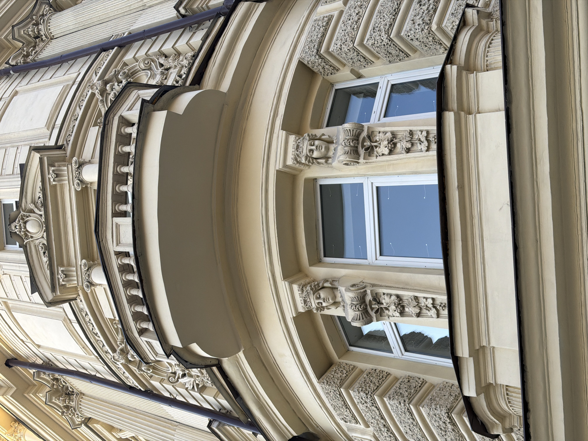 The image shows an upward view of the facade of a building in Moscow, Russia. The architecture features ornate, classical detailing including columns, arches, and decorative stone carving. Windows are visible, reflecting the sky. The color palette is dominated by cream and beige tones.