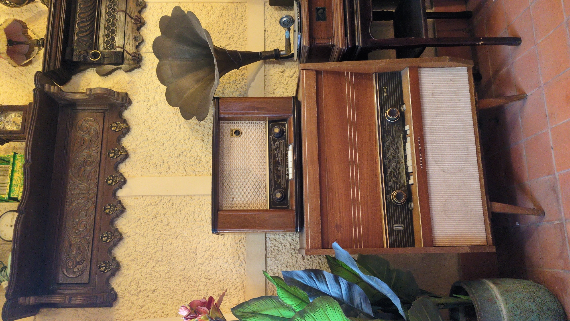 The image depicts a collection of vintage electronics, primarily radios and a gramophone, displayed against a textured yellow wall. The scene appears to be set indoors, possibly in a shop or museum, with terracotta floor tiles visible at the bottom.
Dominating the foreground is a large, wooden console radio with a white fabric speaker grille at the bottom and a complex tuning dial with buttons and knobs above. The brand name "TELEFUNKEN" is visible on the front. Stacked on top of this console is a smaller, similar wooden radio with a more intricate speaker grille. To the right of these radios is a vintage gramophone with a large, metallic horn.
Above the radios, a dark wooden shelf is mounted on the wall, adorned with decorative carvings and small, ornate hooks. On this shelf, several objects are visible, including a clock and a green decorative box. A dark wooden decorative piece with carved details and a metal ring is mounted to the right of the shelf.
To the left of the radios, a large, leafy plant in a green pot adds a touch of natural color to the scene. A single pink flower is also visible on the plant. The overall impression is one of a curated display of historical audio equipment, evoking a sense of nostalgia. The location is not explicitly identifiable as Da Lat, Vietnam, based solely on the visual information presented.