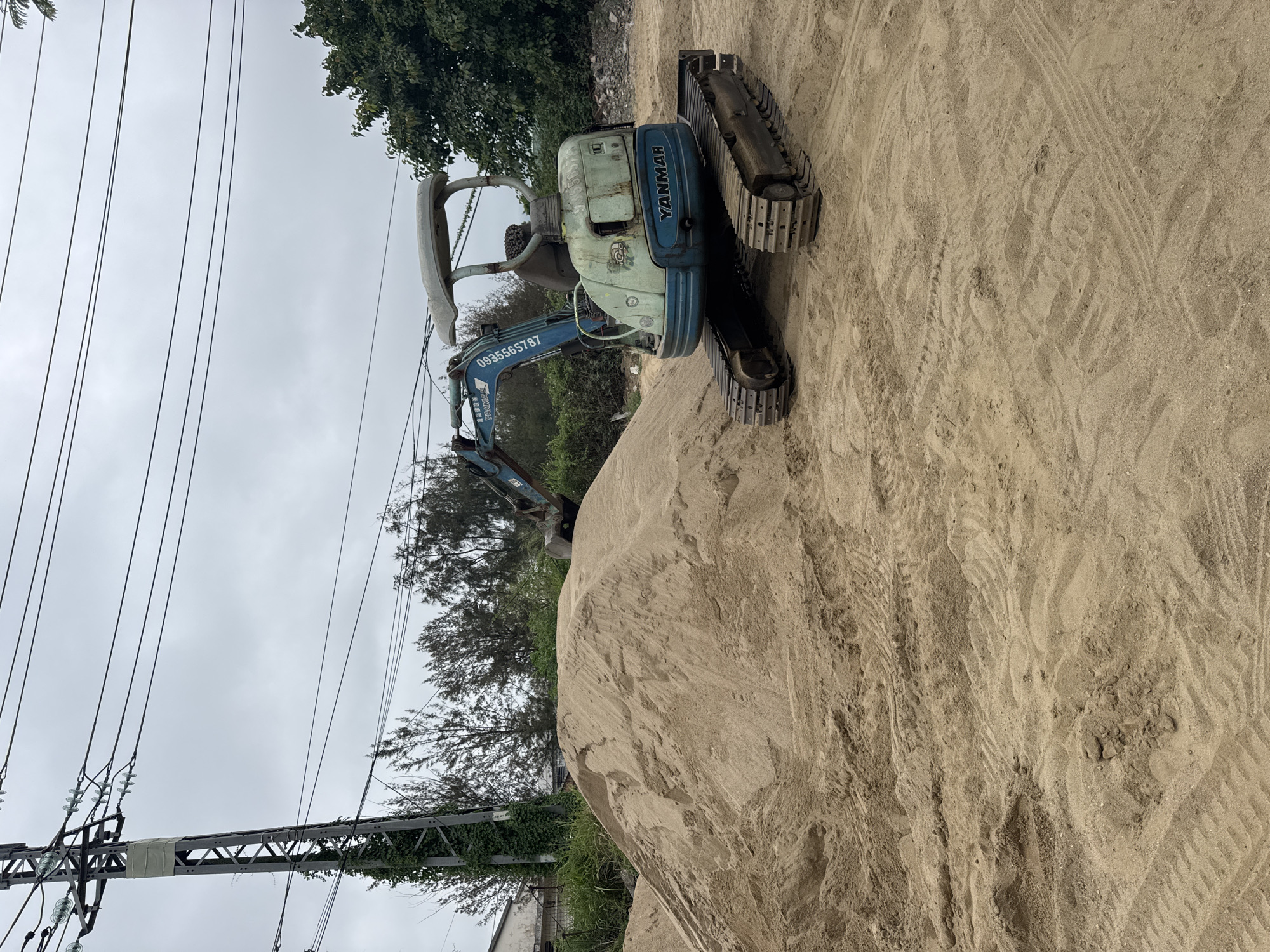 A small, blue-and-white Yanmar excavator sits atop a large pile of sand. The excavator is positioned such that its tracks are visible on the right side of the frame. The machine's arm extends out, but the bucket is obscured by the sand pile. The number "0935565787" is prominently displayed on the arm. Behind the sand pile and excavator, there are trees and foliage. In the distance, more trees and a cloudy sky are visible. In the lower-left corner of the frame, there's a utility pole with numerous power lines. The location is the city of Đà Nẵng, Vietnam.