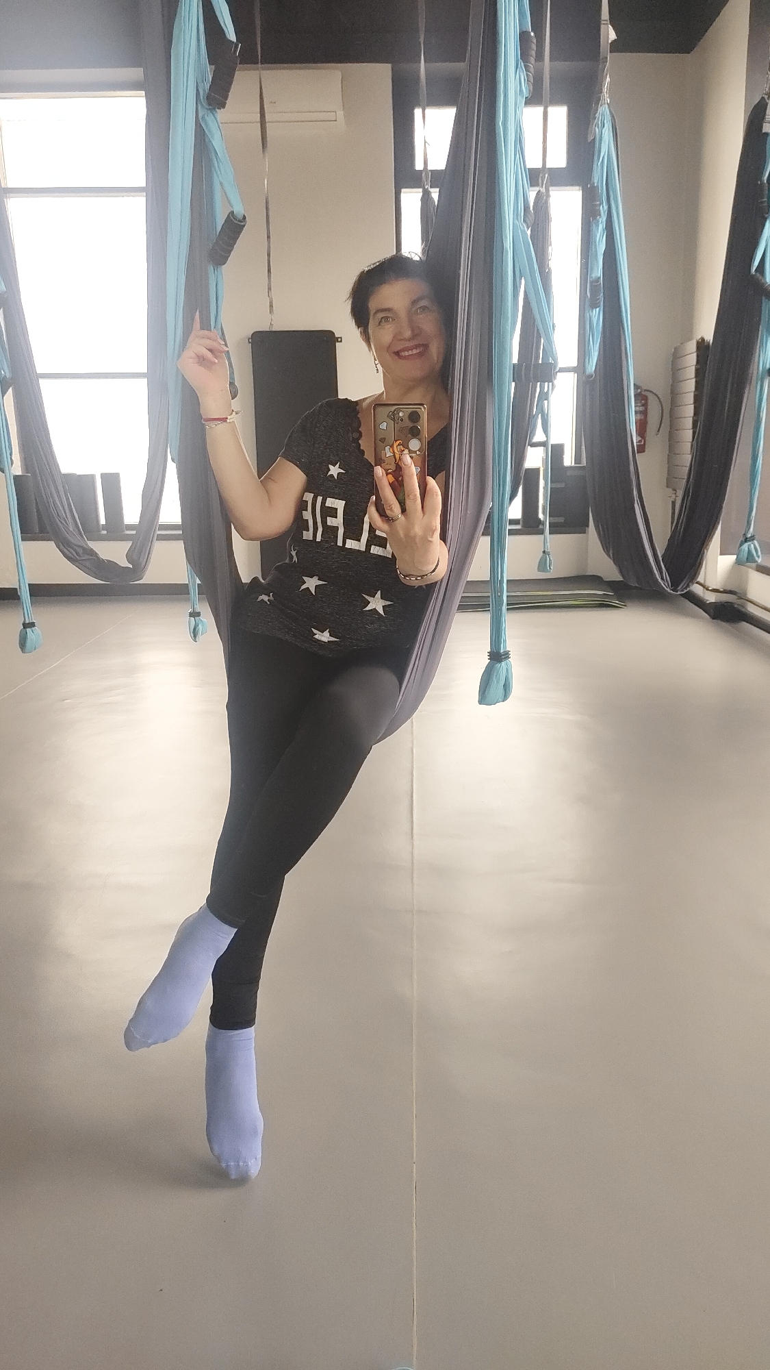 A smiling woman is captured in a selfie taken in an aerial yoga studio in Krasnoyarsk, Russia. She appears to be in her 40s or 50s, with short, dark hair, and is wearing a dark gray t-shirt adorned with white stars and the text "LIVE FREE" (visible in reverse due to the mirror reflection), black leggings, and light blue socks. She is seated comfortably within a dark gray aerial yoga hammock, suspended from the ceiling. Her right hand holds a smartphone with a colorful, patterned case, taking the photo. Her broad smile indicates enjoyment and contentment.
The studio is bright and well-lit, with natural light streaming through large windows on the left side, suggesting it is daytime. Numerous aerial yoga hammocks, in both light blue and dark gray, hang throughout the room, some equipped with handles. The floor is a light-colored, reflective surface, possibly padded, characteristic of a fitness or yoga studio. In the background, reflections show other suspended hammocks, an air conditioner unit on the wall, and a red fire extinguisher on a far wall. The setting appears clean and spacious, designed for aerial fitness activities.