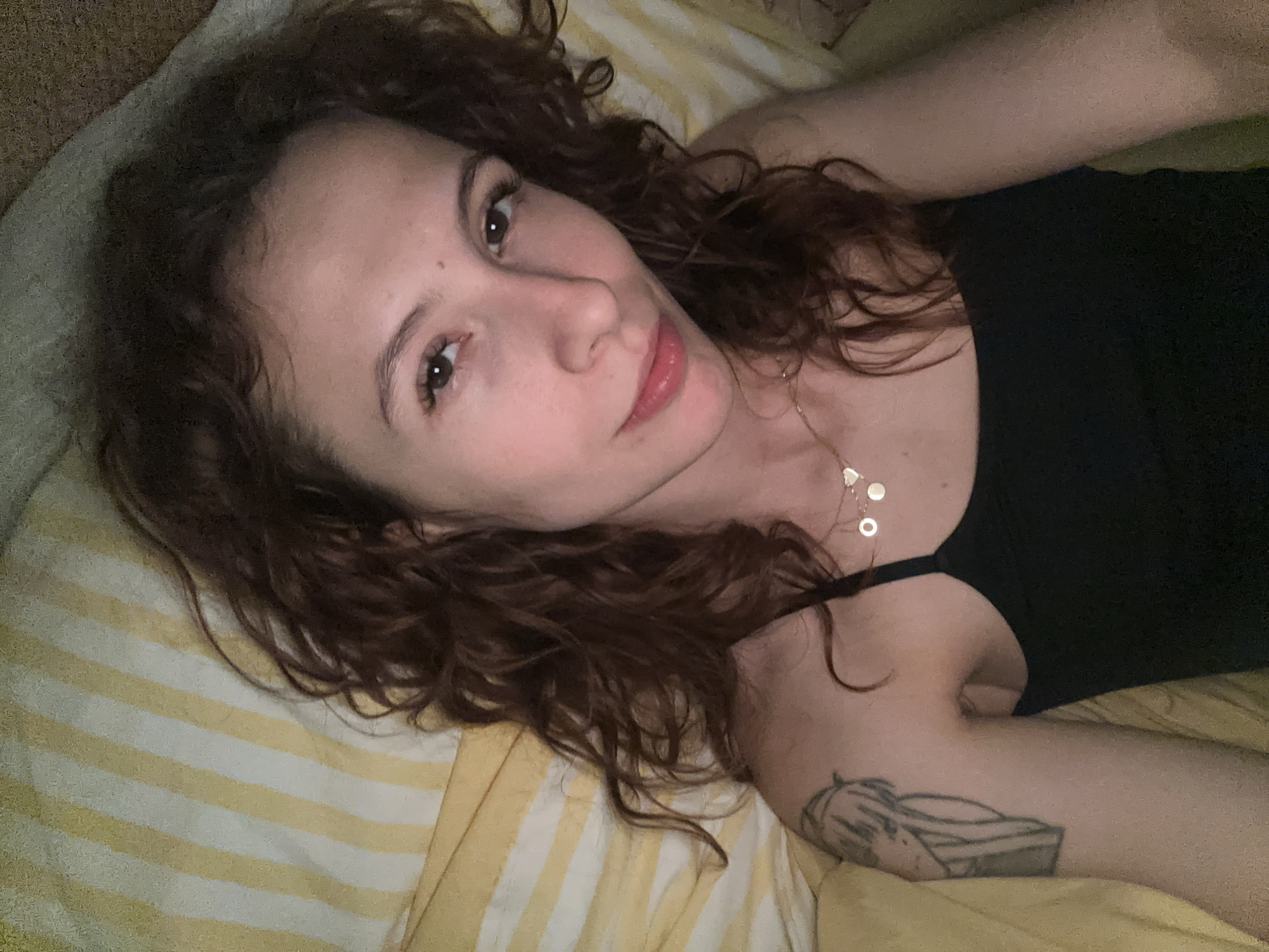 A woman with curly brown hair is lying on a yellow and white striped bed. She is wearing a black tank top and a necklace with circular pendants. She has a tattoo on her right arm. The background includes a plain colored wall or surface. There is no evidence indicating the location is Novi Sad, Serbia.