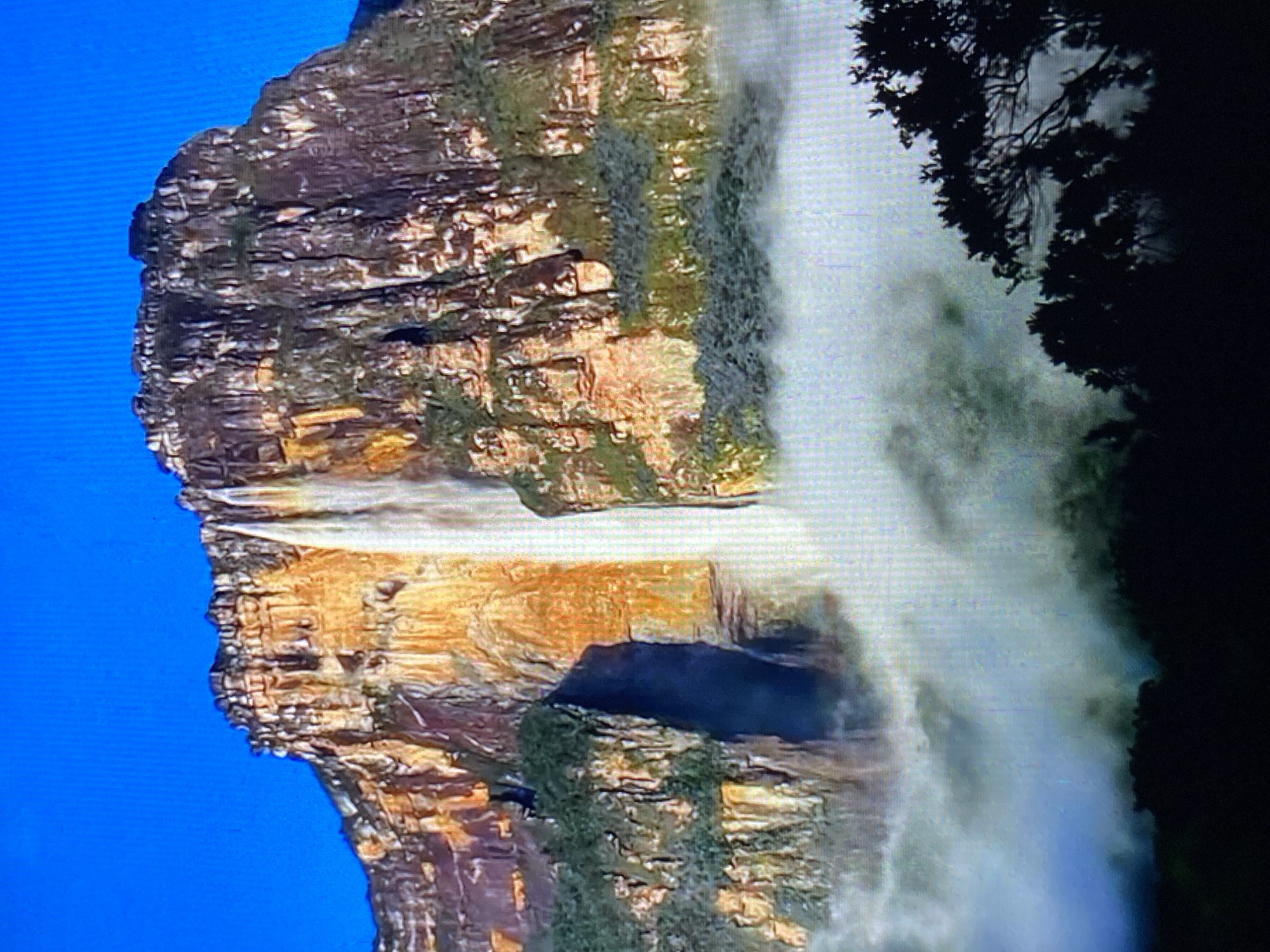 The image shows Angel Falls in Canaima National Park, Venezuela. The falls cascade down a sheer cliff face of layered rock formations into a misty pool. The top of the cliff is rocky, with some vegetation clinging to its surface. A dark outline of trees is visible on the right side of the image. The sky is bright blue.