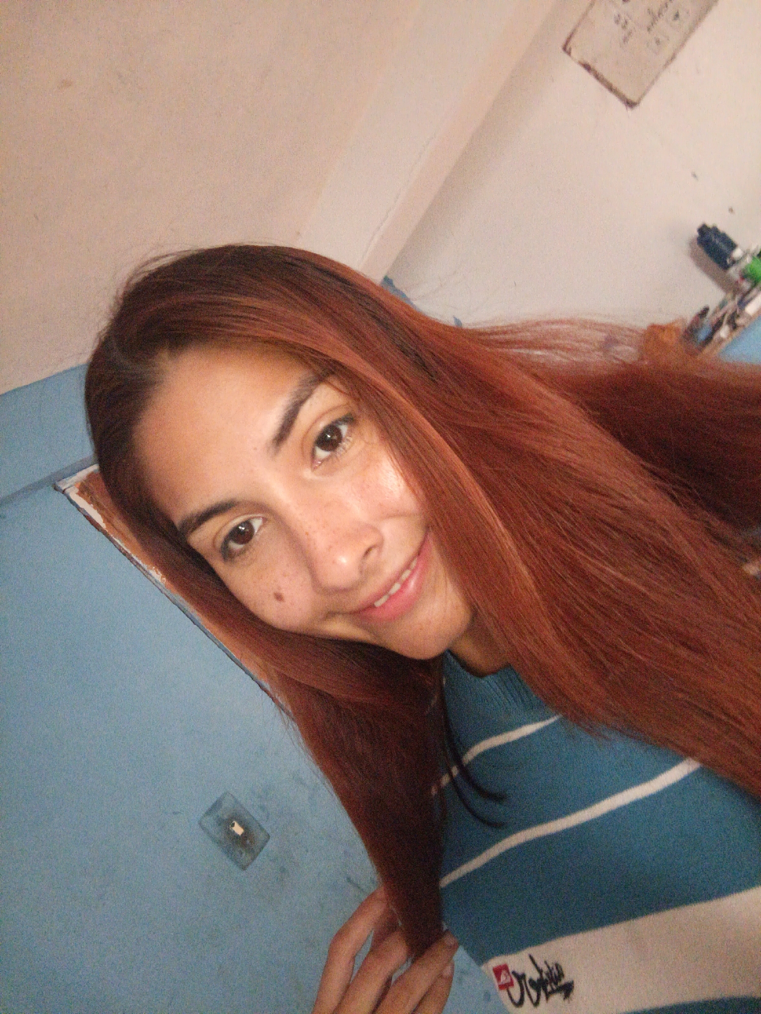 A woman with long, auburn hair smiles into the camera. She has freckles on her face and is wearing a blue and white striped shirt. The background is a light blue wall with a light switch and a white wall with writing on it. The location is Juan Griego, Venezuela.
