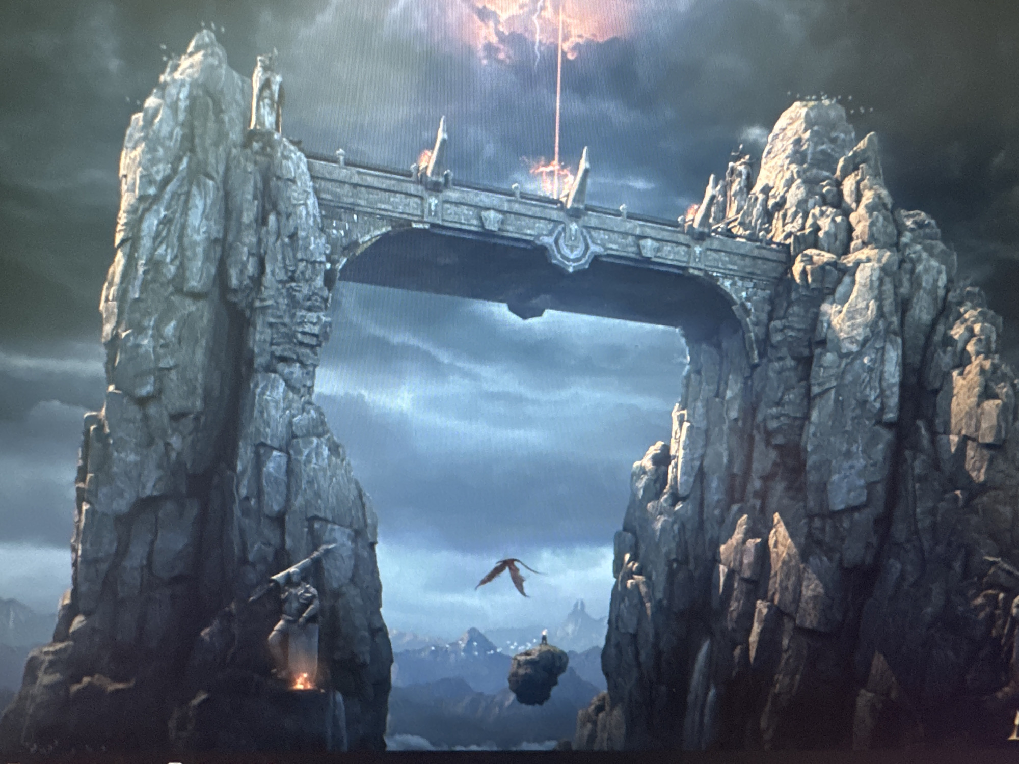 The image depicts a fantastical, mountainous landscape dominated by a large, arched stone bridge connecting two colossal rock formations. The bridge features ornate carvings and is lit by torches on its parapets. Above the bridge, a beam of orange light, possibly lightning, strikes the sky, illuminating dramatic clouds.
In the foreground, on the left rock formation, a warrior-like figure stands by a small fire, holding a spear. In the middle ground, a small, dark dragon with outstretched wings flies between the rock formations. In the far distance, a solitary figure stands on a floating rock amidst a range of mountains. The overall scene is dark and atmospheric, with a focus on dramatic geological formations and mythical elements.
This image does not depict any part of the city of Stepnogorsk city administration, Kazakhstan.