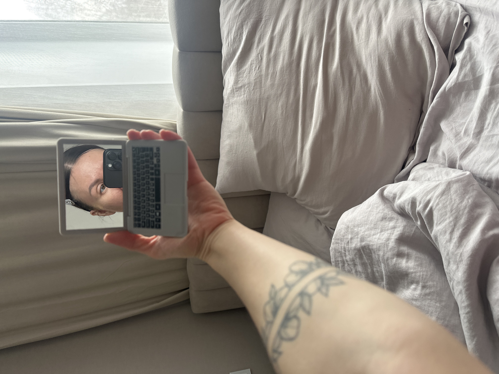 A first-person perspective photograph taken indoors shows an arm with a floral tattoo holding a small, reflective compact. The reflection in the compact reveals a partial view of a person's face, specifically the forehead, eye, and a phone held close to the face. The background includes a bed with light gray bedding and a headboard. The lighting suggests daylight. The location is not discernible from the provided image and thus cannot be placed in the city of Votkinsk, Russia.