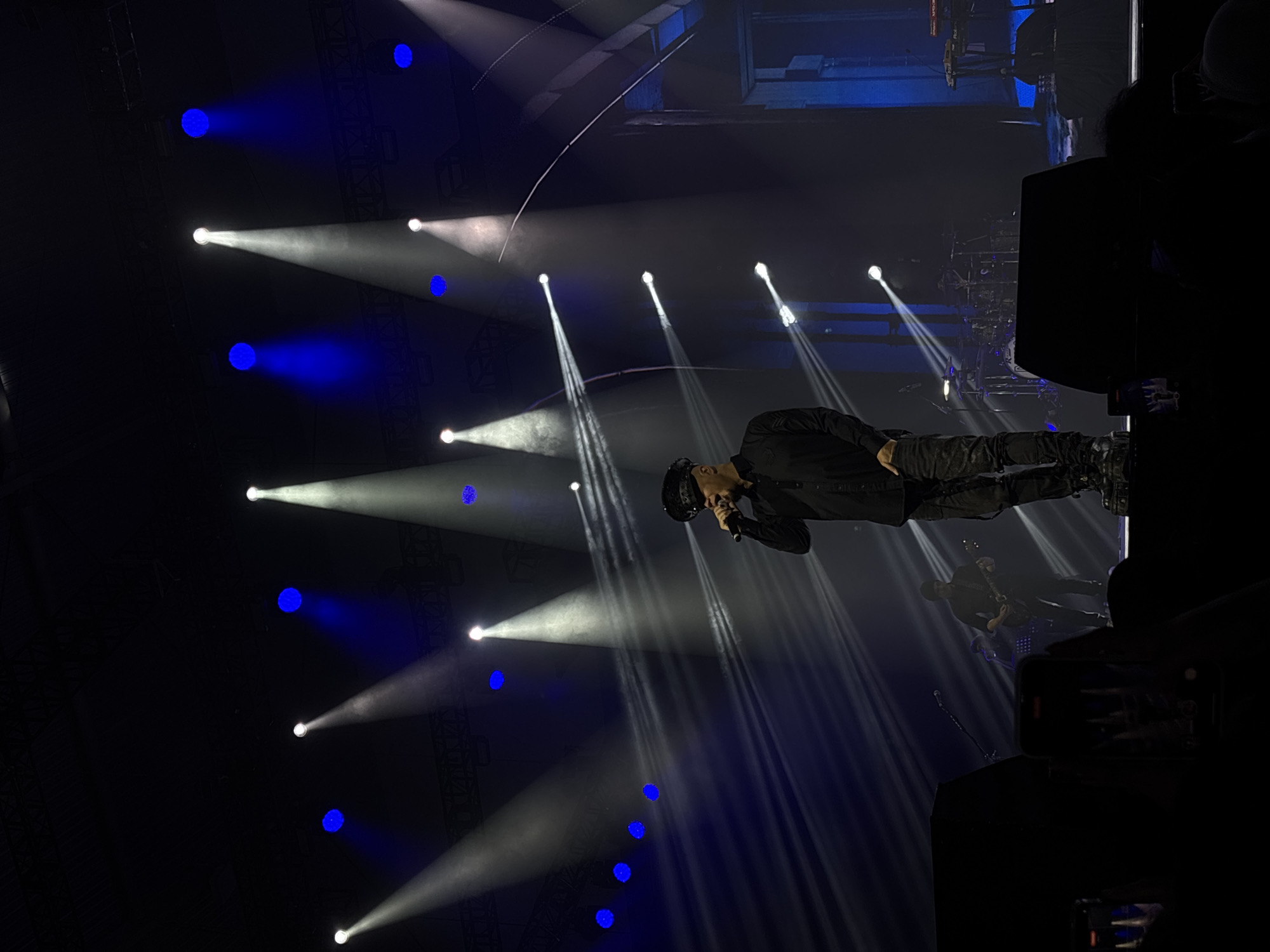 A performer stands on a stage illuminated by spotlights in Pagedangan, Indonesia. The performer is wearing dark clothing and holding a microphone. Behind the performer, there are other performers and musical instruments visible. Stage lighting in the form of white spotlights and blue stage lights are visible and directed toward the performers on the stage. Audio equipment and possibly audience members are visible in the darkness surrounding the stage.