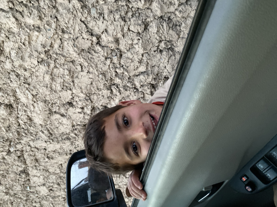 A young male, appearing approximately 7-10 years old, with short dark hair and brown eyes, smiles directly at the viewer. He is positioned outside a vehicle, leaning into an open window, with his left hand resting on the top edge of the light-grey car door. He wears a light-toned shirt, possibly with a reddish collar visible beneath. The car's side mirror is visible in the upper left, reflecting a partial, indistinct view of the vehicle's interior. In the lower left foreground, power window and door lock controls are discernible on the car's interior door panel. The background consists of a rough, textured wall, consistent with unplastered mud or adobe construction, characteristic of structures in Kandahar, Afghanistan.