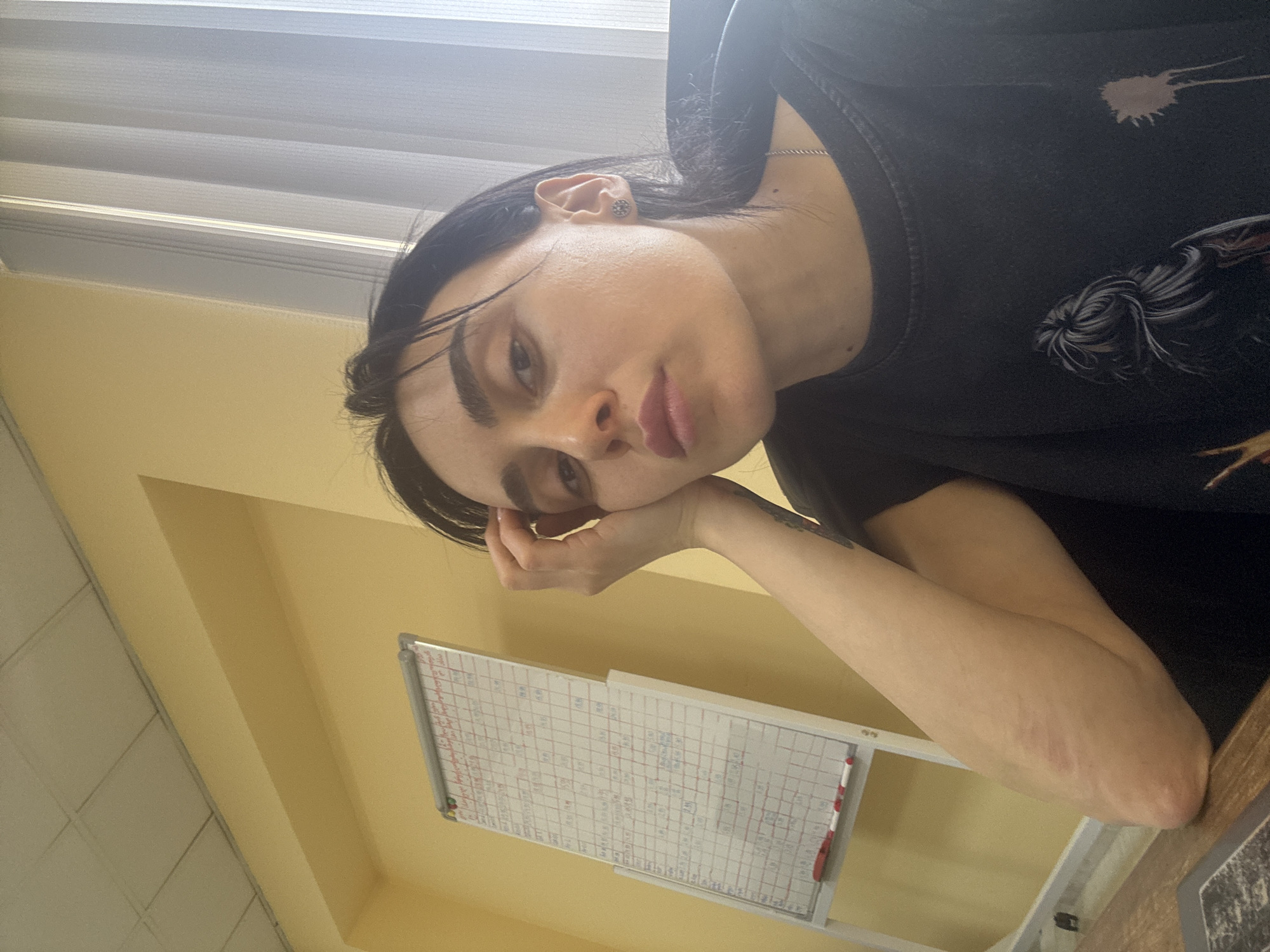 The image shows a person with fair skin, dark hair, and noticeable eyebrows posing with their head resting on their hand. They are wearing a black t-shirt with a graphic design and a silver necklace. A whiteboard with a grid pattern is mounted on the wall behind them. The background includes a yellow-painted wall, and a window with white blinds. The scene appears to be indoors, potentially in an office or workspace. The location in the city of Tbilisi, Georgia, is not explicitly provided in the image.