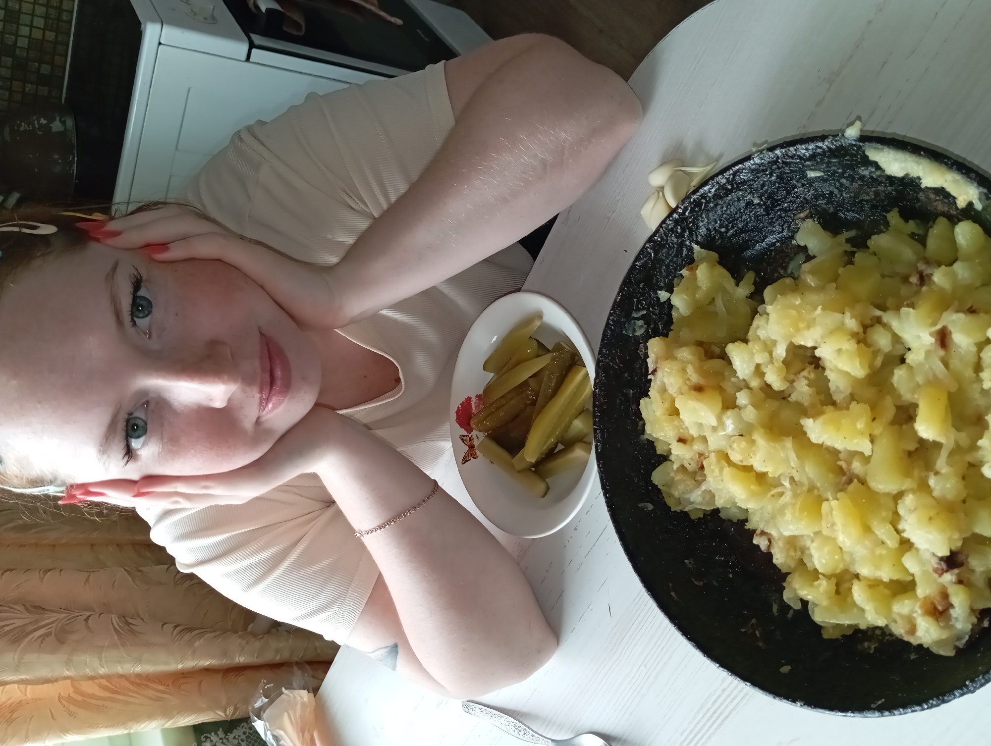 A fair-skinned, red-haired woman sits at a white table with a cast-iron pan of fried potatoes and a plate of pickles. Her hands rest on her cheeks. The photograph appears to be taken in a residential kitchen; a stove is partially visible in the background. There is no indication that the photo was taken specifically in Yurgamysh, Russia.