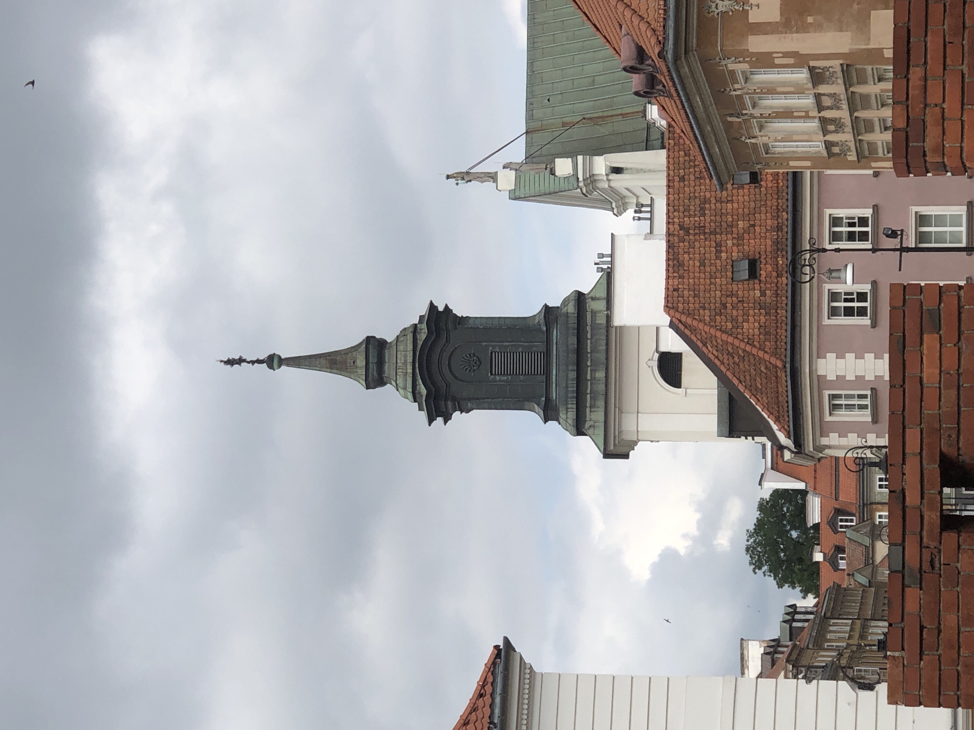 The image depicts a view of the Old Town Market Square in Warsaw, Poland, under a cloudy sky. The central focus is the spire of a church, likely St. John's Archcathedral, with its dark green, tiered structure topped by a cross. To the right of the spire, a green-tiled roof of another building is visible, adorned with two statues on its peak. In the foreground, the red brick wall of a fortress or old building runs across the bottom of the frame. Behind the wall, various buildings with red-tiled roofs and cream-colored facades are seen, interspersed with windows of different sizes and designs. One building, painted a pale pink, has a distinctive lamp post in front of it. In the upper right portion of the sky, a single bird is captured in flight.