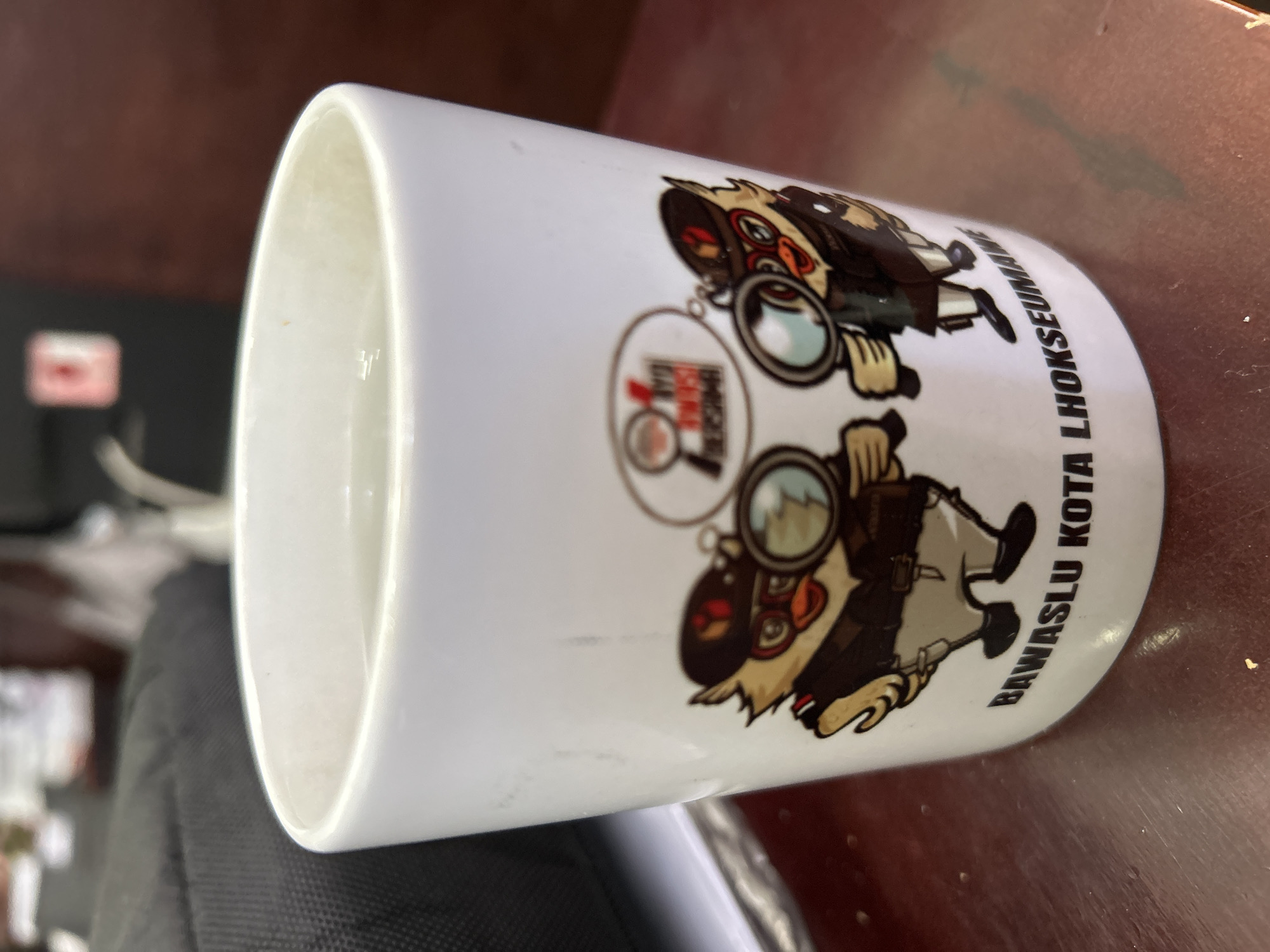 A white mug with a cartoon detective otter character is displayed on a brown surface. The mug features an emblem depicting the same otter character holding a magnifying glass inside a circular logo, with text that reads "BAWASLU KOTA LHOKSEUMAWE" around the lower part of the mug. The background shows blurred elements.
