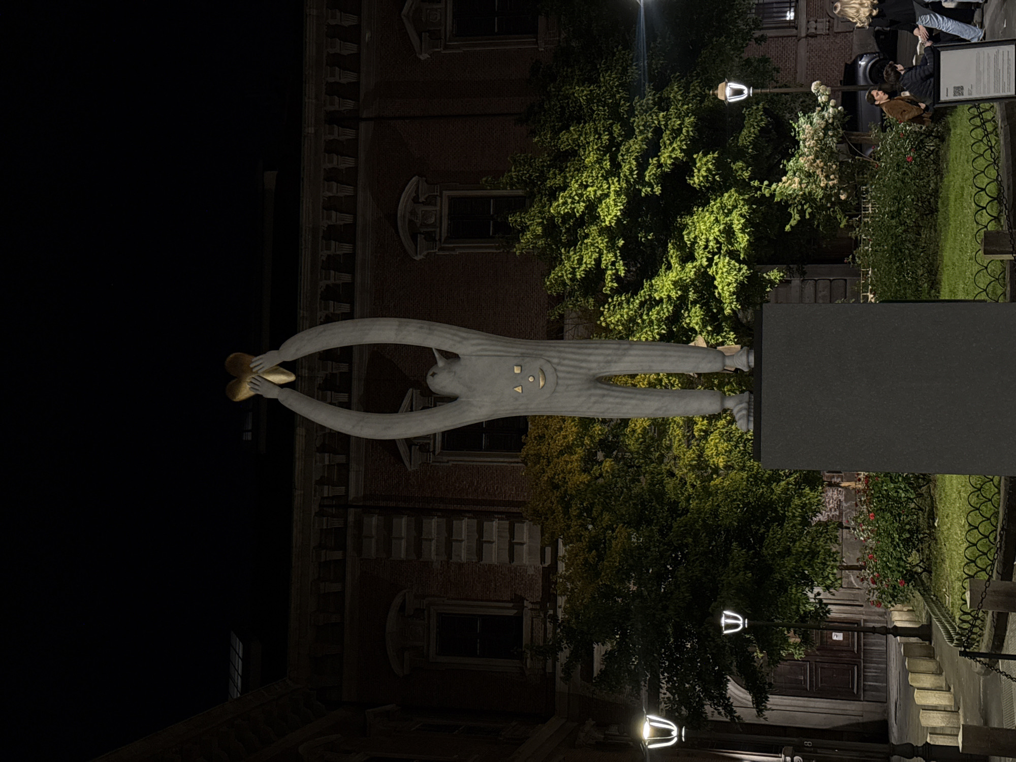 This night photograph, taken in Milan, Italy, depicts an outdoor urban scene featuring a prominent sculpture, illuminated foliage, and a small group of people. The image is presented vertically, with the description assuming a correctly upright orientation.
The central subject is a tall, light-colored sculpture of a stylized humanoid figure with an elongated body. Its arms stretch upwards, holding a golden, heart-shaped or clover-like object. The figure's face features simple, smiling details: two eyes, a nose, and an upward-curving mouth. It stands on a dark, rectangular pedestal.
In the mid-ground to the right, several individuals are gathered casually. One person with blonde hair, dressed in a black top and light-colored pants, stands near another person in a brown jacket who appears to be seated or leaning. They seem to be conversing or observing their surroundings. A black car is partially visible behind them.
The background features a multi-story brick building with distinct European architectural elements, including decorative cornices and a mix of arched and rectangular windows. Lush green trees and bushes, brightly illuminated by artificial lights, punctuate the dark night setting. Patches of white and red flowers are visible within the foliage.
The scene is lit by several modern-designed street lamps. A dark display board with white text and a QR code is positioned near the gathered people, likely providing information about the artwork or location. A dark metal fence with decorative patterns separates parts of the area. The overall ambiance suggests a serene public space in a historic city, with the illuminated sculpture serving as a focal point.