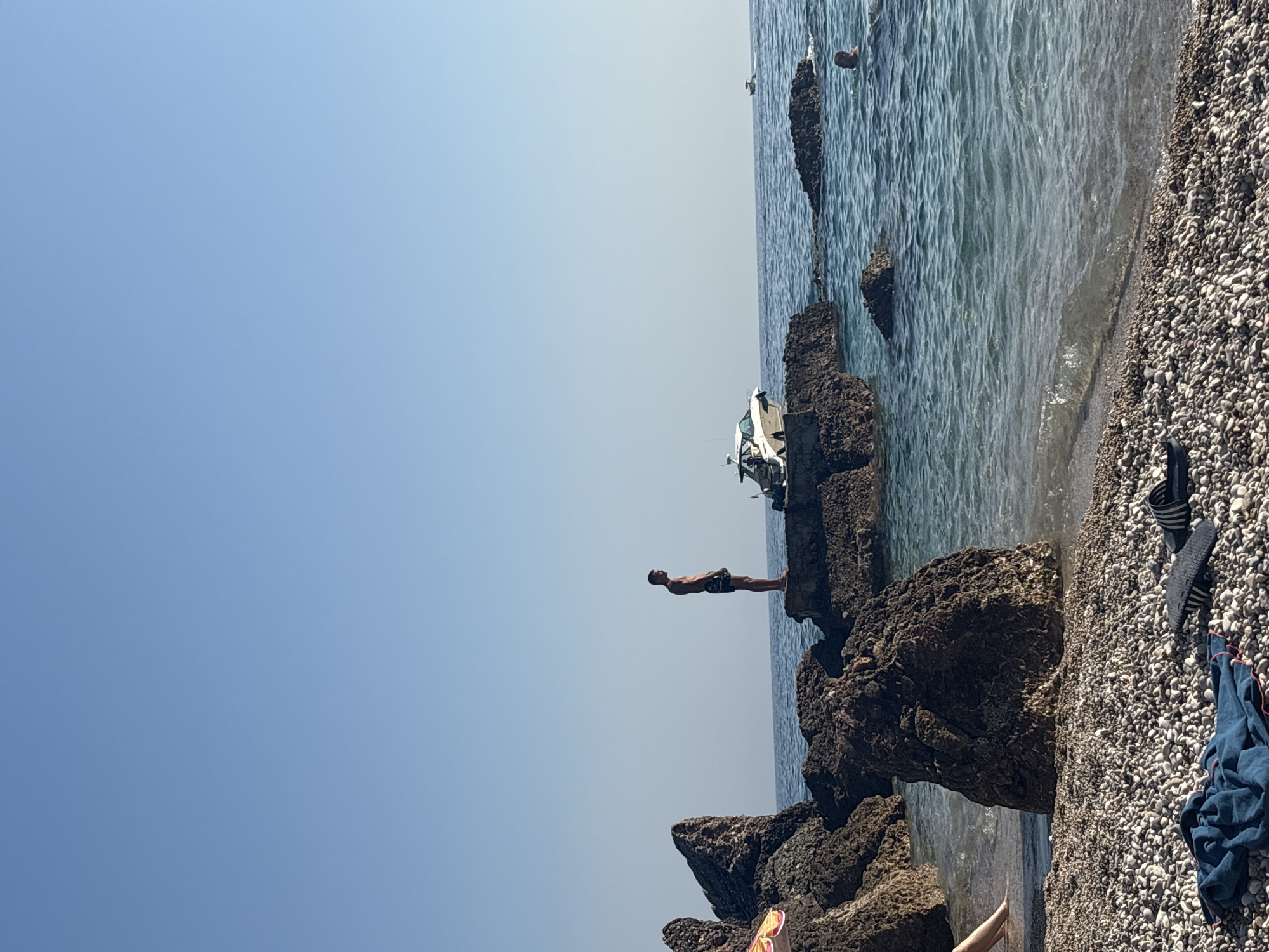 A man stands on a rock formation at the edge of the Adriatic Sea in Drobnići, Montenegro. The man, wearing dark swimming trunks, faces away from the camera, looking out at the water. Behind him, a white motorboat is docked near the rocks. The sea is calm and blue, with a clear sky overhead. In the foreground, a pebble beach is visible, with a blue towel and sandals lying on the stones. Another person is seen swimming in the water further out. The scene depicts a peaceful day at the beach.