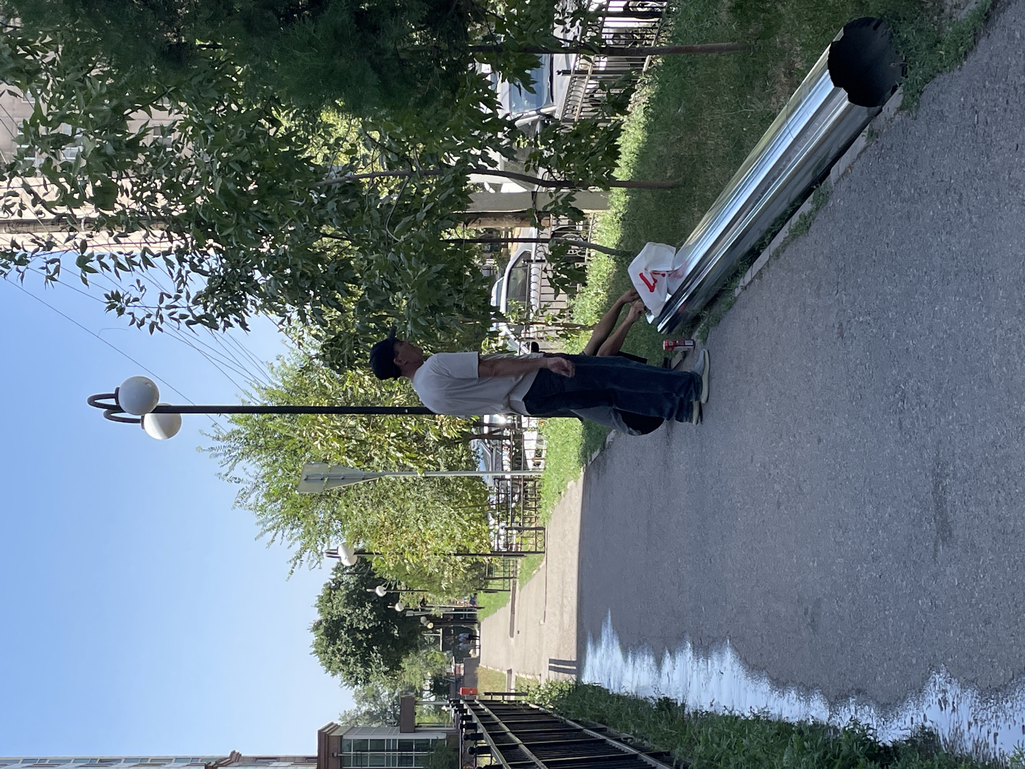 The image depicts a street scene in Almaty, Kazakhstan, under a clear blue sky. A man wearing a white t-shirt and dark pants stands on a sidewalk, looking down at a large, metallic pipe lying on the grass next to the sidewalk. Another person, whose face is not visible, is crouched down, interacting with the pipe and a white plastic bag with red lettering that is on top of it. A red can is also visible near the pipe. The sidewalk runs parallel to a black metal fence, beyond which is a grassy area and trees. In the background, there are cars parked along the street and buildings. Streetlights with round, white lamps are visible along the sidewalk. The overall scene suggests outdoor maintenance or construction work.