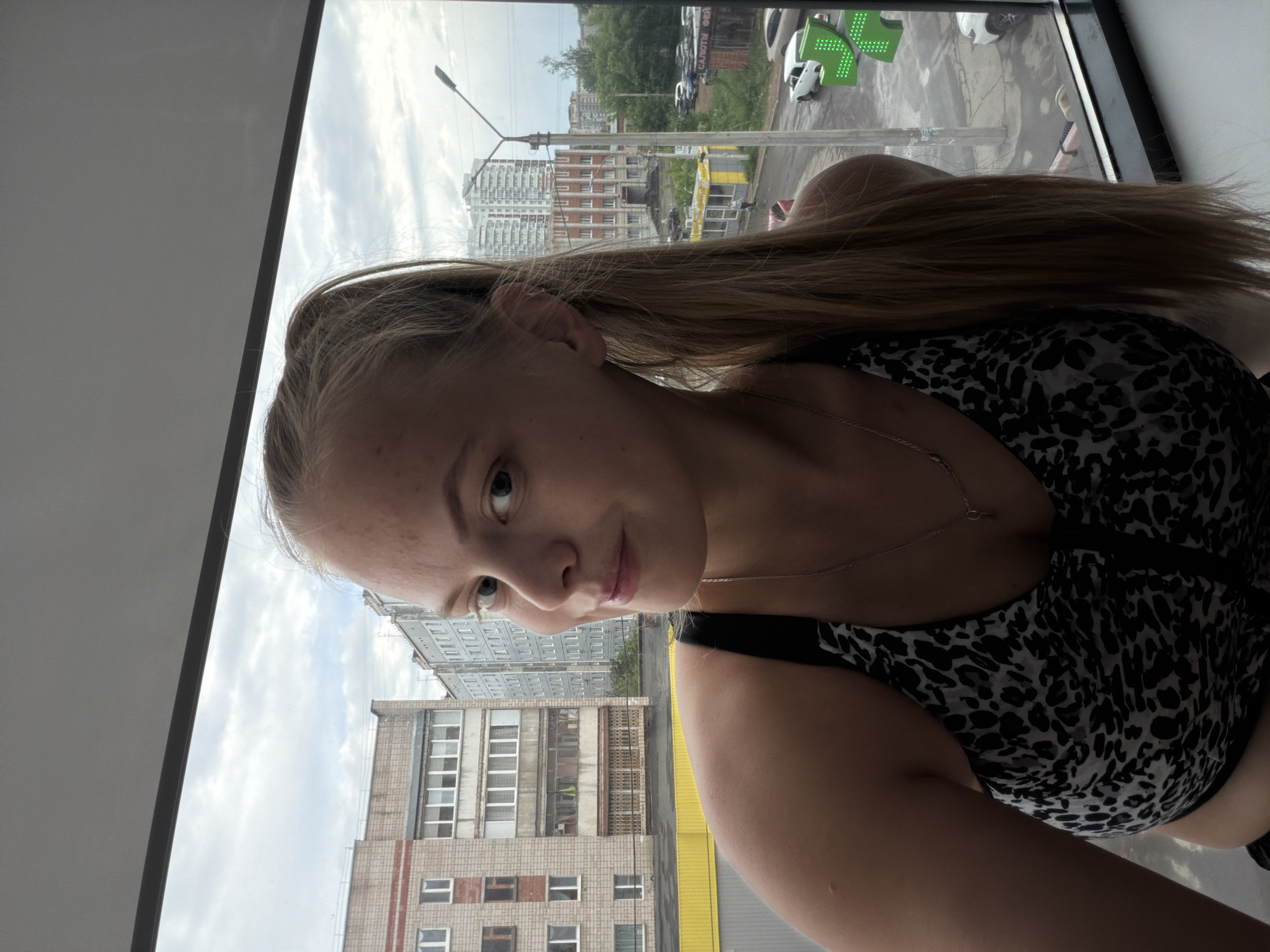 A young, blonde woman with her hair in a ponytail poses close to a window overlooking an urban scene. She is wearing a black and white patterned top and a delicate necklace. Through the window, one sees a cityscape with buildings of various heights and construction styles. There's a pharmacy indicated by a green cross. A street, possibly a road junction, is visible, with cars and what appears to be a pedestrian crossing. Based on the architecture and identifiable signage, the location is Izhevsk, Russia. The weather appears overcast with a mix of clouds and some blue sky.