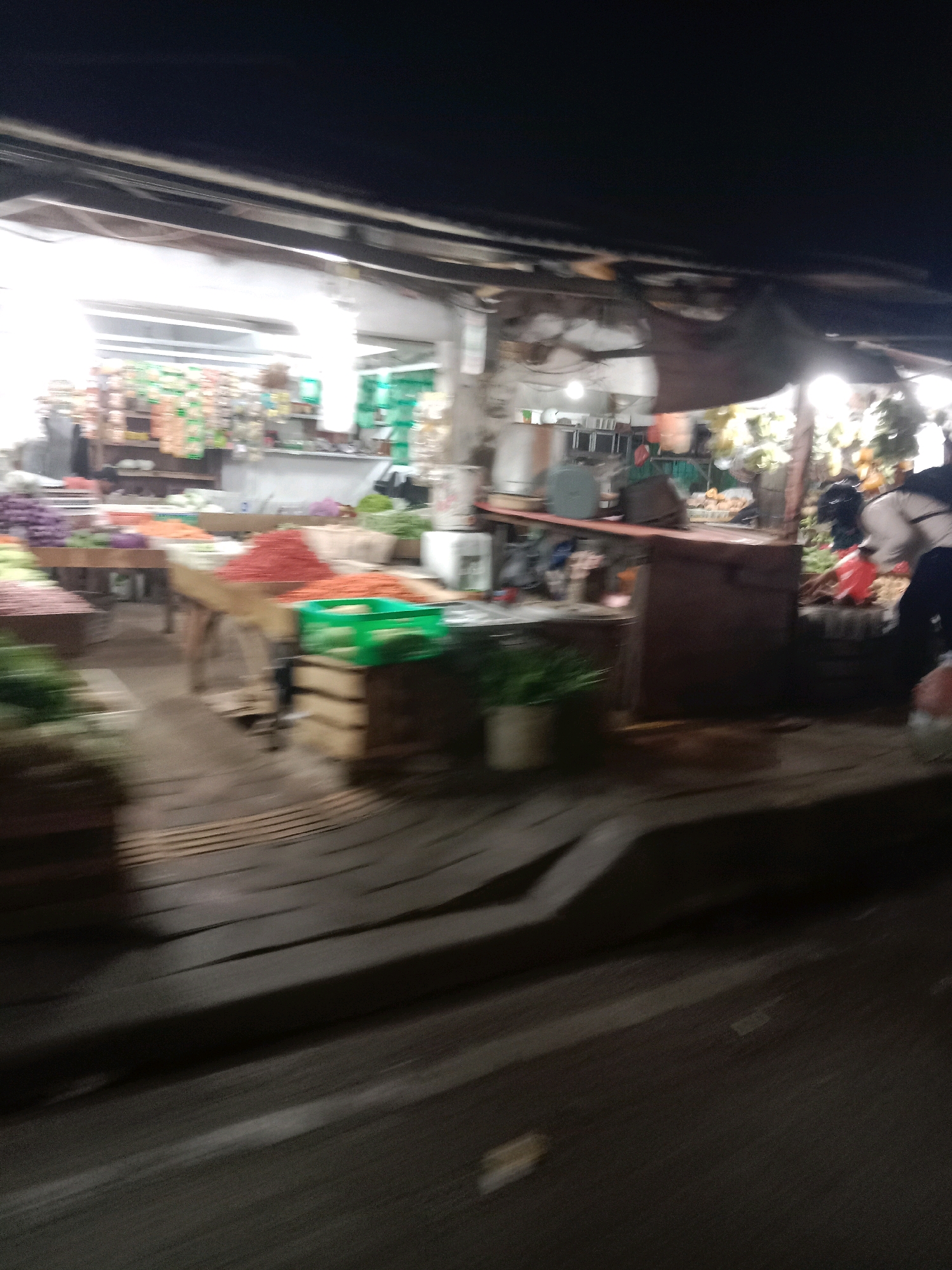 The image shows an outdoor market stall in South Tangerang, Indonesia, at night. The market appears to be a produce market with various fruits and vegetables displayed on wooden tables and in crates. The lighting suggests it is after dark, possibly with artificial lights illuminating the stalls. There are some people visible in the background, their forms blurred due to motion or low light conditions. The overall scene depicts a bustling, active marketplace.