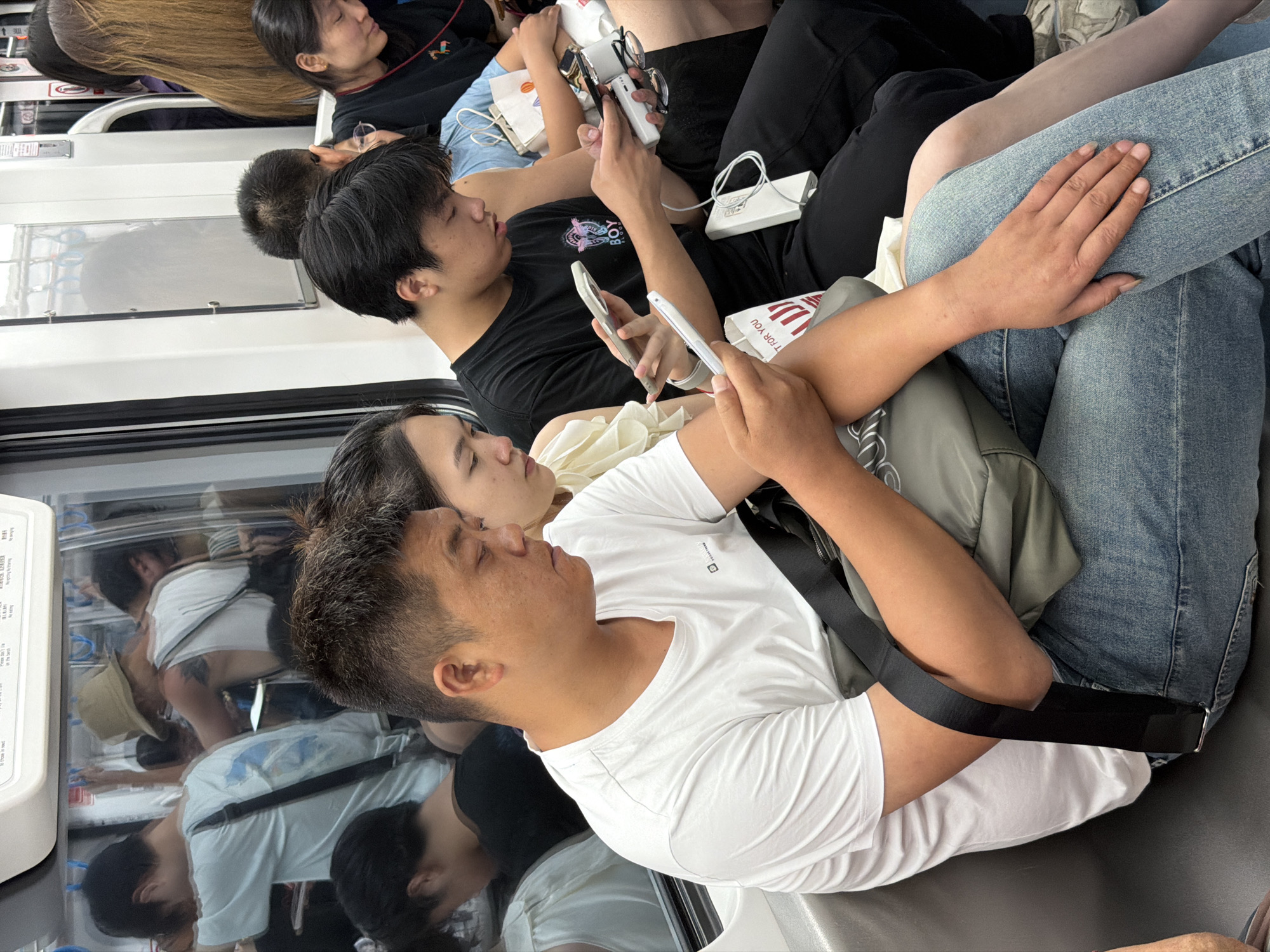 The image displays the interior of a crowded subway train car. In the foreground, a man with short, dark hair and a tanned complexion is seated, wearing a white short-sleeved t-shirt and light blue jeans. A dark olive-green cross-body bag is strapped across his chest. He is focused on a white smartphone held in his left hand. To his right, a woman with long dark hair, wearing a light-colored ruffled top, is partially visible, also looking downwards. Adjacent to her, a younger man with short, dark hair, dressed in a black t-shirt with a graphic design featuring text and an illustration, is actively using two smartphones; a white one in his left hand and a darker one in his right. A white power bank and charging cables are visible near his right hand. A white paper bag with red text, partially reading "T FOR YOU," rests on his lap. To the far right, a woman with long, straight, light brown hair, wearing a black top with colorful embroidery, is holding a dark smartphone in her left hand and a white device, possibly another power bank, in her right.
Through the train's window, reflections of several standing passengers are visible. These include a person wearing a beige bucket hat and a white tank top with a tattoo on their left upper arm, and another person in a light blue t-shirt. Overhead grab handles are visible in the reflections. The train's interior features grey seating, white wall panels, and silver overhead handrails. Partially visible signs above the window display text in an Asian script and English, including phrases such as "to those in need" and "Please don't lean on the doors." The scene indicates heavy passenger traffic, with most individuals engrossed in their mobile devices.