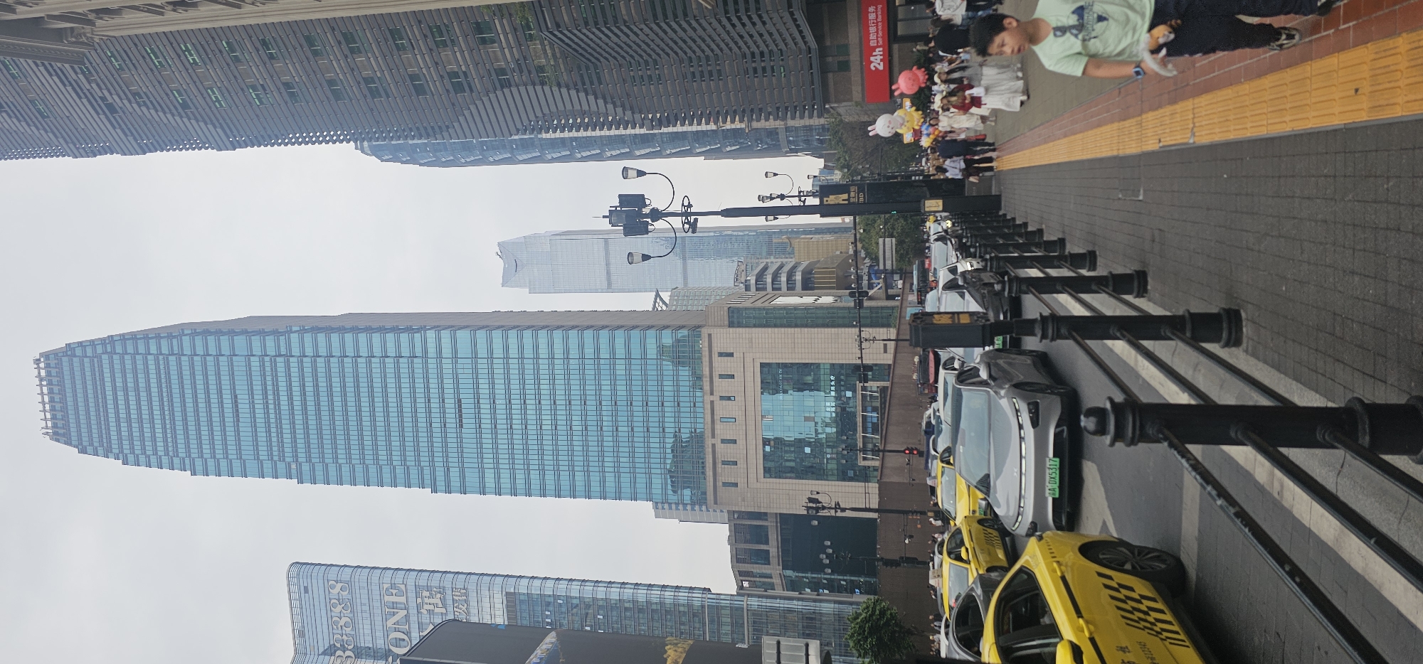 The image depicts a street scene in the Yuzhong District, China. Tall buildings line the street, including a prominent skyscraper with reflective blue windows. A row of taxis, predominantly yellow, is visible along the street, with a gray car interspersed among them. A metal railing runs along the edge of the sidewalk, separating it from the street. On the sidewalk, there is a pedestrian area where people are walking, some carrying balloons. A young boy is visible on the right side of the image, walking along the sidewalk. The sky is overcast.