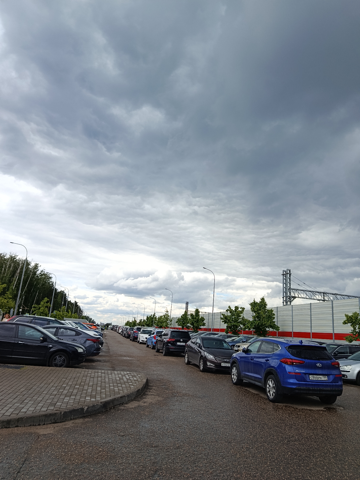 A long line of cars is parked on a paved road. The sky is overcast with dark, heavy clouds. On the left side of the road, there is a line of trees and streetlights. On the right side, there is a large white structure with a red trim, possibly a sound barrier, and some more trees. The cars are of various colors and models, and they are parked bumper to bumper, suggesting a busy area or event. The road surface appears wet in places, indicating recent rain.