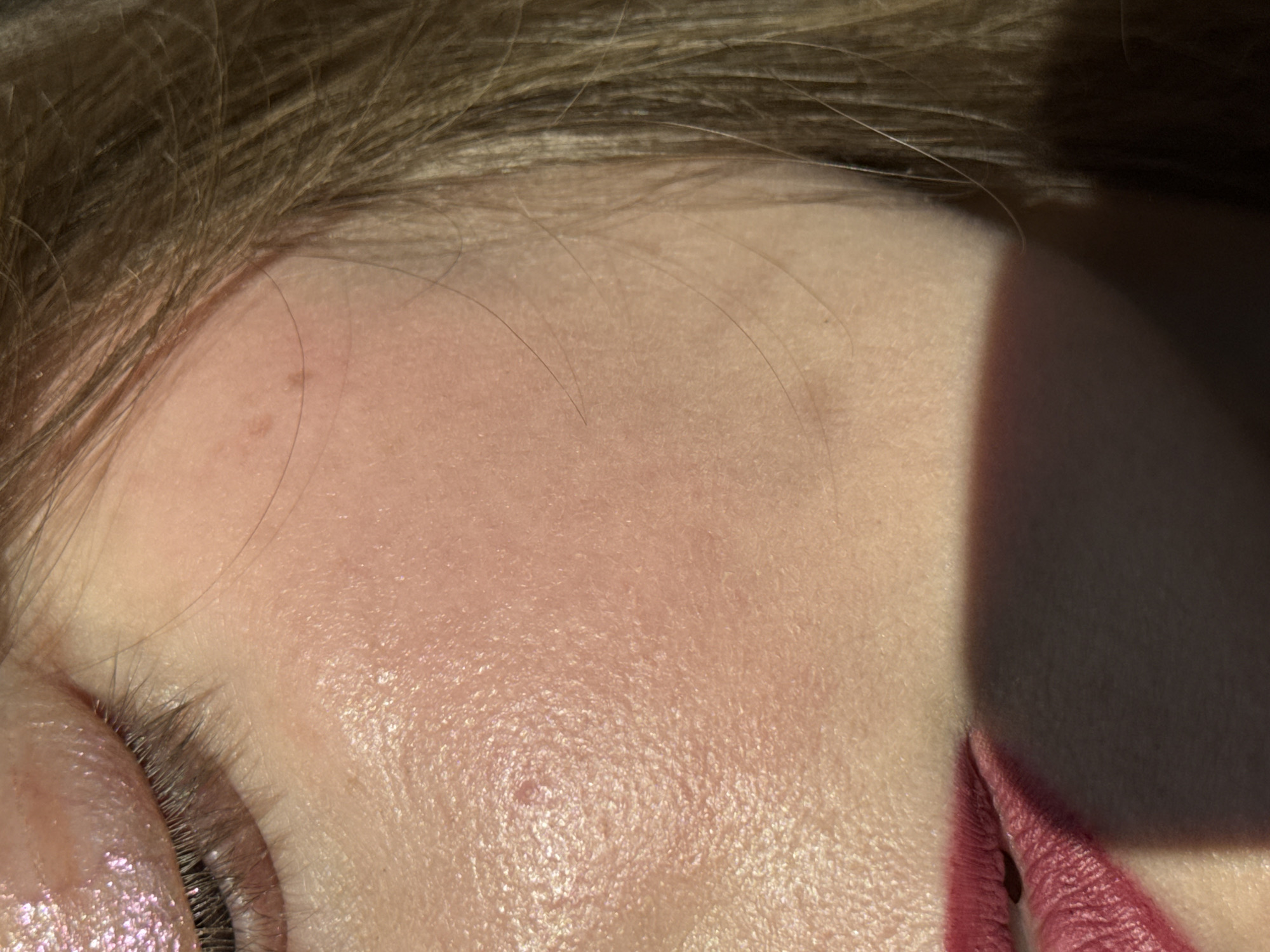 The image shows a close-up of a person's face, specifically the forehead, eye, and part of the lips. The skin appears to be fair. The eye is made up with a light eyeshadow and dark eyelashes. The lips are visible in the lower right corner and are wearing a dark pink lipstick. The upper left corner shows the person's hairline with light brown hair. A shadow falls across the right side of the face. There is no information to deduce the location as Izhevsk, Russia.