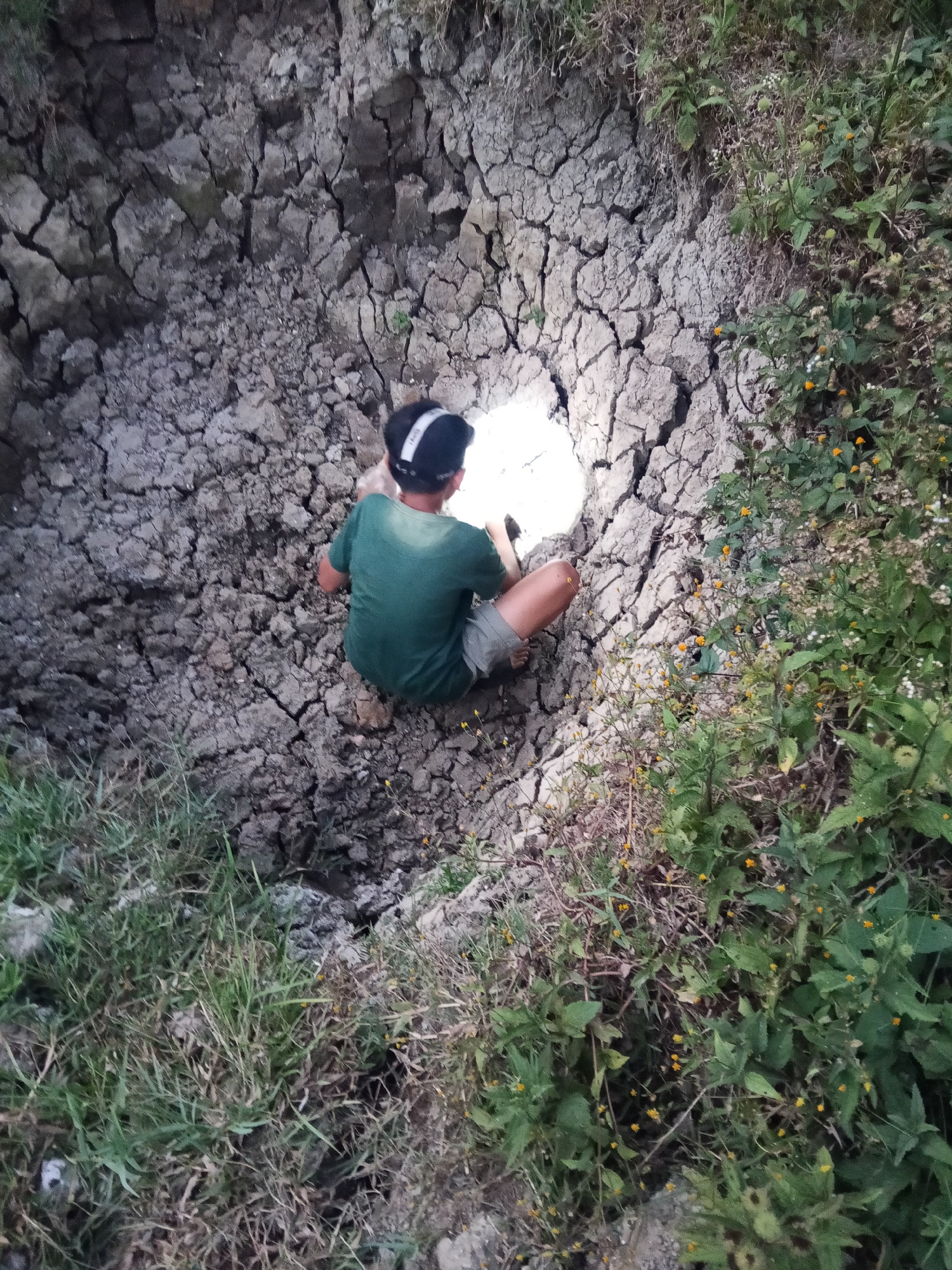 The image shows a young person, seen from behind, wearing a green t-shirt and shorts, crouching in a cracked, dry earth pit. The person is holding a light source, illuminating a small area of the pit. The pit is surrounded by dry, cracked earth and some overgrown vegetation with small yellow flowers on the right side. The scene appears to be an outdoor environment, possibly in a rural or undeveloped area. The location is specified as the city of Makale, Indonesia.