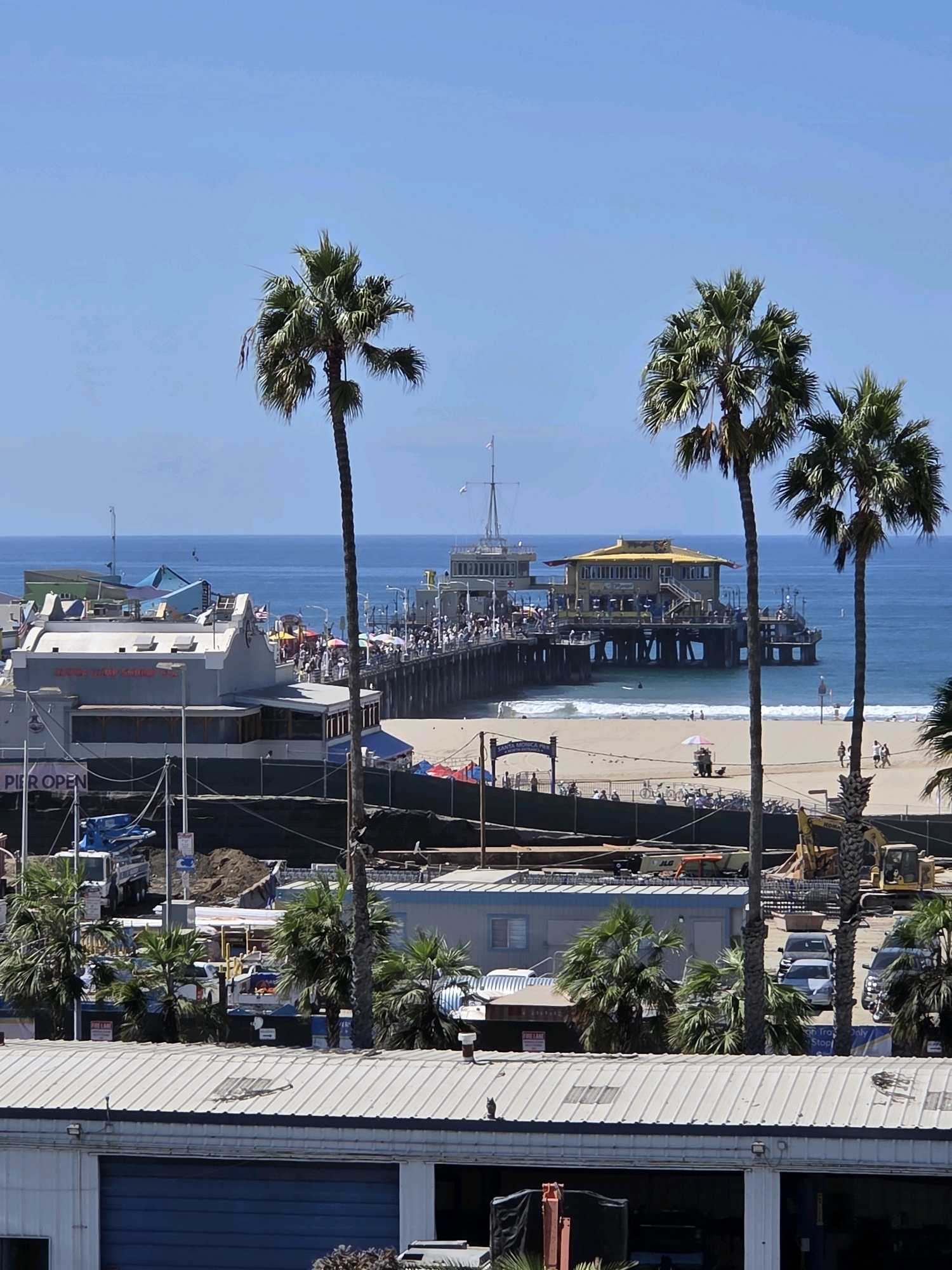 This is a full, eye-level, outdoor, daytime, eye-level medium shot of the Santa Monica Pier in Santa Monica, California, USA. The scene is dominated by palm trees in the foreground, with a wide view of the pier extending into the ocean. The sky is clear and blue, with no clouds visible. The ocean is calm and blue, with gentle waves lapping at the shore.
The pier is a popular tourist destination and is bustling with activity. People can be seen walking, shopping, and dining at the various establishments on the pier. The iconic Ferris wheel and roller coaster are visible, along with a variety of food vendors and shops. The beach is also crowded with people enjoying the sun and sand. The scene is vibrant and lively, capturing the essence of a typical day at the Santa Monica Pier.
In the foreground, several palm trees are visible, their fronds swaying gently in the breeze. Behind the palm trees, the pier stretches out into the ocean, a vibrant hub of activity. The architecture of the pier is a mix of classic and modern, with a variety of colorful buildings and attractions. The beach is a stretch of golden sand, dotted with umbrellas and beach towels.
In the middle ground, the ocean extends as far as the eye can see, a brilliant expanse of blue. The waves are gentle, creating a serene and peaceful atmosphere. The horizon is crisp and clear, with no signs of pollution or smog.
The overall impression of the scene is one of a lively and exciting tourist destination. The Santa Monica Pier is a place where people come to relax, have fun, and create lasting memories.
