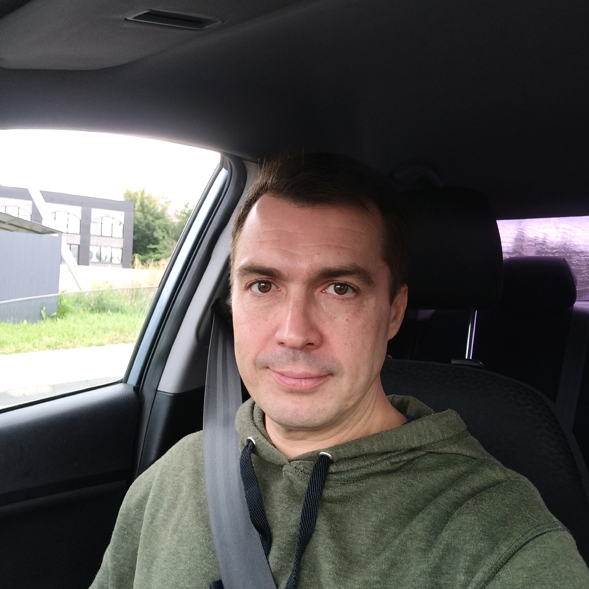 A man is pictured from the chest up, sitting in the driver's seat of a car. He is looking directly at the camera with a slight smile. He is wearing a green hooded sweatshirt with drawstrings. A gray seatbelt is visible across his chest. Through the car window, a building and some greenery can be seen, suggesting an outdoor setting. The interior of the car, including the headrest and part of the dashboard, is also visible. There is no information to confirm this image is located in Nizhny Novgorod, Russia.