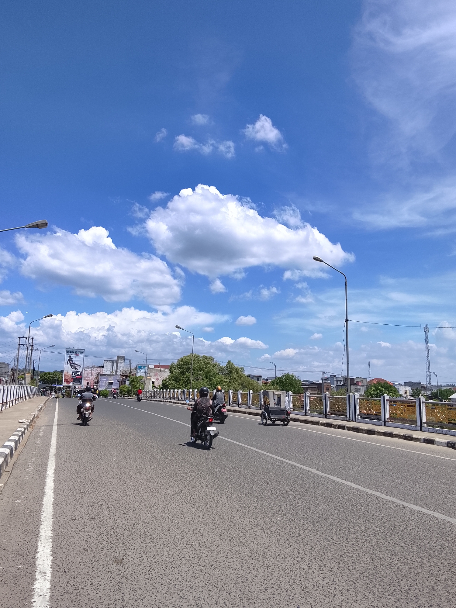 The image depicts a view of the Krueng Cunda bridge in Lhokseumawe, Indonesia, on a bright, sunny day. The sky is a vibrant blue, dotted with fluffy white clouds. The bridge itself is a multi-lane road with a distinctive ornamental railing along the sides. A white line delineates the lanes of traffic.
Several individuals are visible riding motorcycles, traveling in the same direction. One motorcycle carries a passenger. A three-wheeled vehicle, likely a becak, is also present on the road. On the side of the road, lampposts are erected. Buildings and trees can be seen in the background, with a telecommunications tower visible on the far right. A billboard advertising "NO FEAR NO STORY" is positioned to the left. The overall scene suggests a typical day of activity on this city bridge.