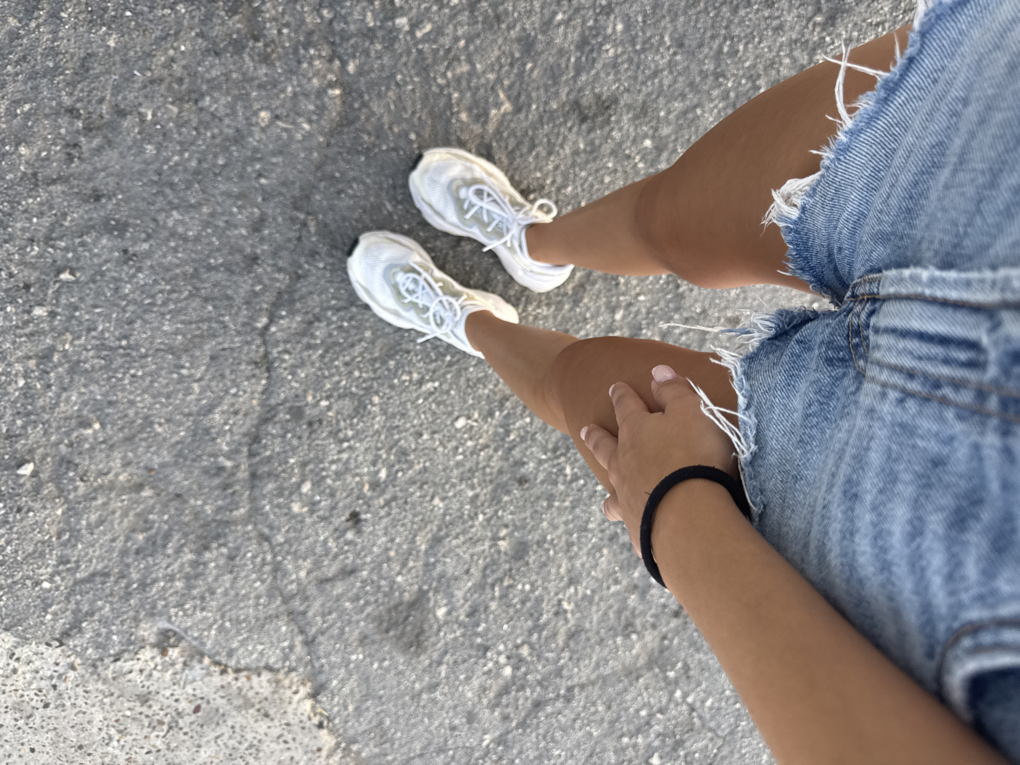 The image is a first-person perspective shot of a person's legs and feet standing on a cracked asphalt surface. The person is wearing distressed denim shorts and white athletic shoes with white laces. One hand, with painted nails, is resting on the thigh. The scene appears to be outdoors, possibly a street or pathway, in Cimișlia, Moldova. The lighting suggests it is daytime.