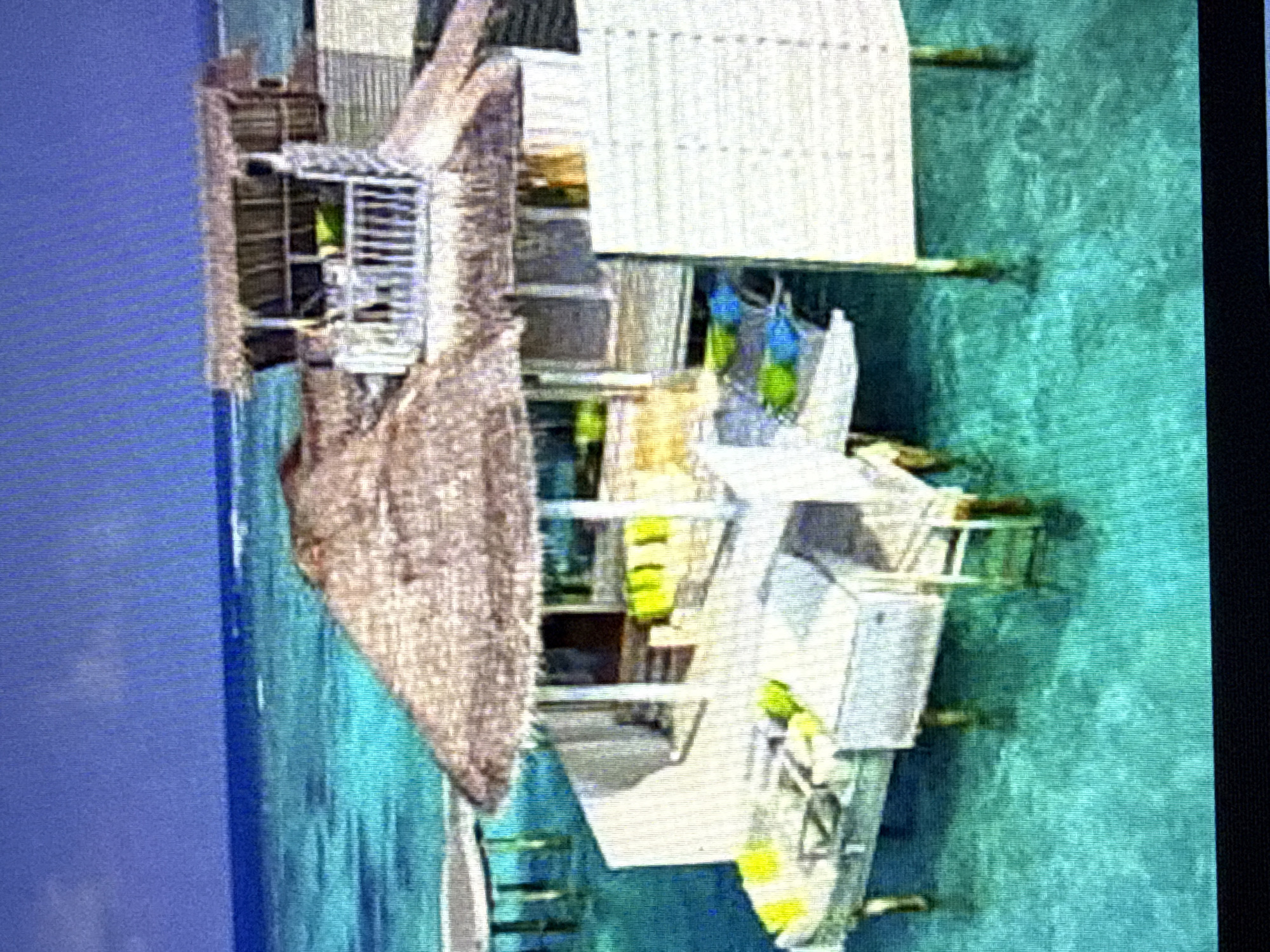 A luxurious overwater bungalow is situated in a clear, turquoise ocean. The bungalow features thatched roofs, white railings, and open-air living spaces furnished with vibrant green and blue cushions. Various platforms and walkways connect different sections of the structure, all supported by wooden stilts in the water. The water is crystal clear, revealing the submerged stilts and the sandy ocean floor in places. The sky above is a deep blue, with a faint horizon line visible. The scene evokes a sense of tropical paradise and relaxation.