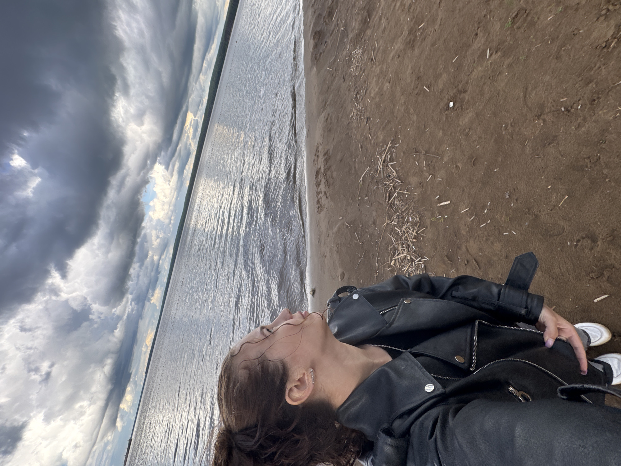 The image, oriented correctly, features a woman in the foreground on a sandy or muddy shoreline. She is seen from the chest up, with her head tilted upwards, looking towards the sky with a contemplative or serene expression. Her dark brown, curly hair appears slightly damp, with a few strands clinging to her face. She is wearing a black leather jacket with silver zippers and snaps, and white sneakers are visible at the bottom of the frame. A small silver earring adorns her left ear.
The setting is a large body of water, likely a lake or wide river, stretching into the distance. The water's surface is rippled, reflecting the light from the sky. A distant, dark treeline marks the opposite shore. The sky is dramatic and heavily overcast with large, dark gray clouds, though some patches of brighter, possibly blue, sky are visible, indicating dynamic weather conditions. The shoreline is composed of dark brown sand or mud, with scattered debris like small sticks and dried grass. No specific activity beyond posing for the photograph is evident. The scene suggests a daytime setting, possibly late afternoon, given the cloud cover and light. The location is contextualized as Chaikovsky, Russia, which aligns with the depiction of a large river or lake. No visible text is present in the image.