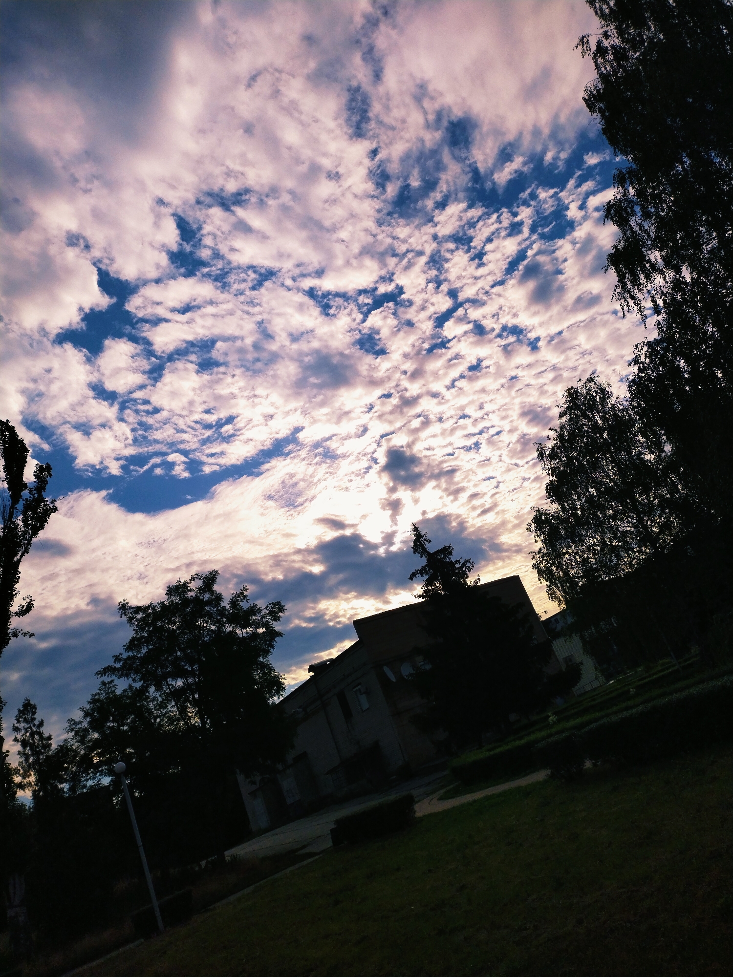 The image is a low-angle shot of a partially cloudy sky over a street or park area in Horishni Plavni, Ukraine. The sky is filled with a mixture of dark and light clouds, with patches of blue sky visible. The sun appears to be setting or rising, casting a warm glow through the clouds. In the foreground, there are silhouetted trees and the top of a building. A street lamp is also visible in the lower left portion of the image. The scene suggests an urban or suburban environment during twilight.