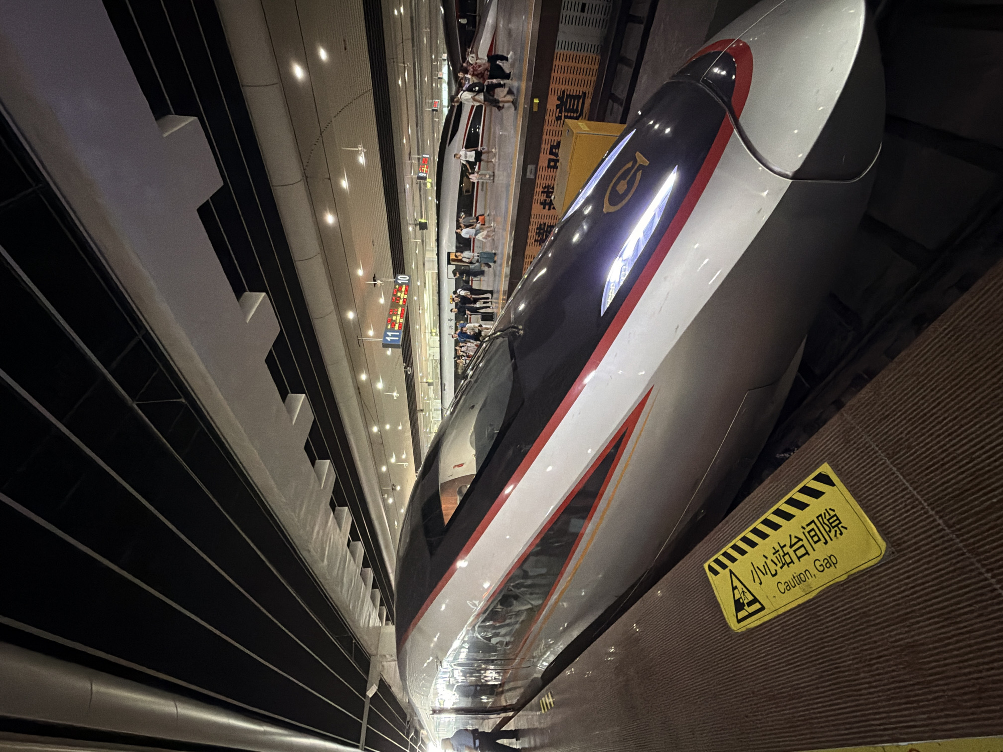 The image captures a scene at a train station in Panyu District, China. In the foreground, the sleek, modern exterior of a high-speed train is prominently displayed, its silver, white, and red livery reflecting the station's lighting. A yellow warning sign with black text and graphics, reading "Caution, Gap" in English and Chinese, is affixed to the textured surface of the train platform.
In the mid-ground, passengers are visible on the platform, some with luggage, preparing to board or disembark from another high-speed train parked alongside. The station's infrastructure, including overhead lighting and signage indicating platform numbers "11" and "10," is visible. The environment appears to be an indoor station with a high ceiling supported by a dark, angular structure. The atmosphere suggests a busy transportation hub during operational hours.