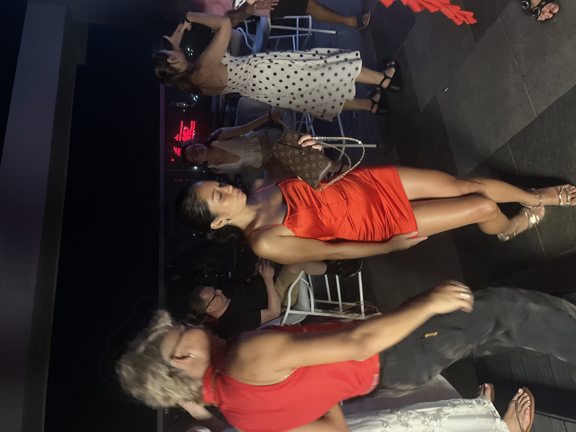 This is a medium-shot, eye-level, candid photograph taken at night, likely at a rooftop venue in Da Nang, Vietnam. The scene depicts several people enjoying themselves, with some dancing and others sitting.
In the foreground, a woman in a bright red, strappy mini-dress is captured mid-dance, her body angled towards the left of the frame. She holds a designer clutch bag with a gold chain. To her left, a person with blonde, spiked hair and wearing a red sleeveless top and dark pants is also in motion, with their back largely to the camera. They appear to be facing towards the left of the frame.
In the mid-ground, a man wearing glasses and a black t-shirt sits on a white chair, looking down at his phone. Behind him, another woman in a white top with a plunging neckline stands, her gaze directed towards the left. Further to the right, a woman in a white dress with black polka dots is visible, also in motion, with her right arm raised. A person in a pink shirt is partially visible on the far right.
The environment appears to be an outdoor space with dark flooring composed of large, square tiles. In the background, a red neon sign with indistinct lettering is visible, suggesting an urban setting. The lighting is dim, with some artificial lights illuminating the subjects and creating shadows on the floor. The overall atmosphere suggests a lively social gathering or party.