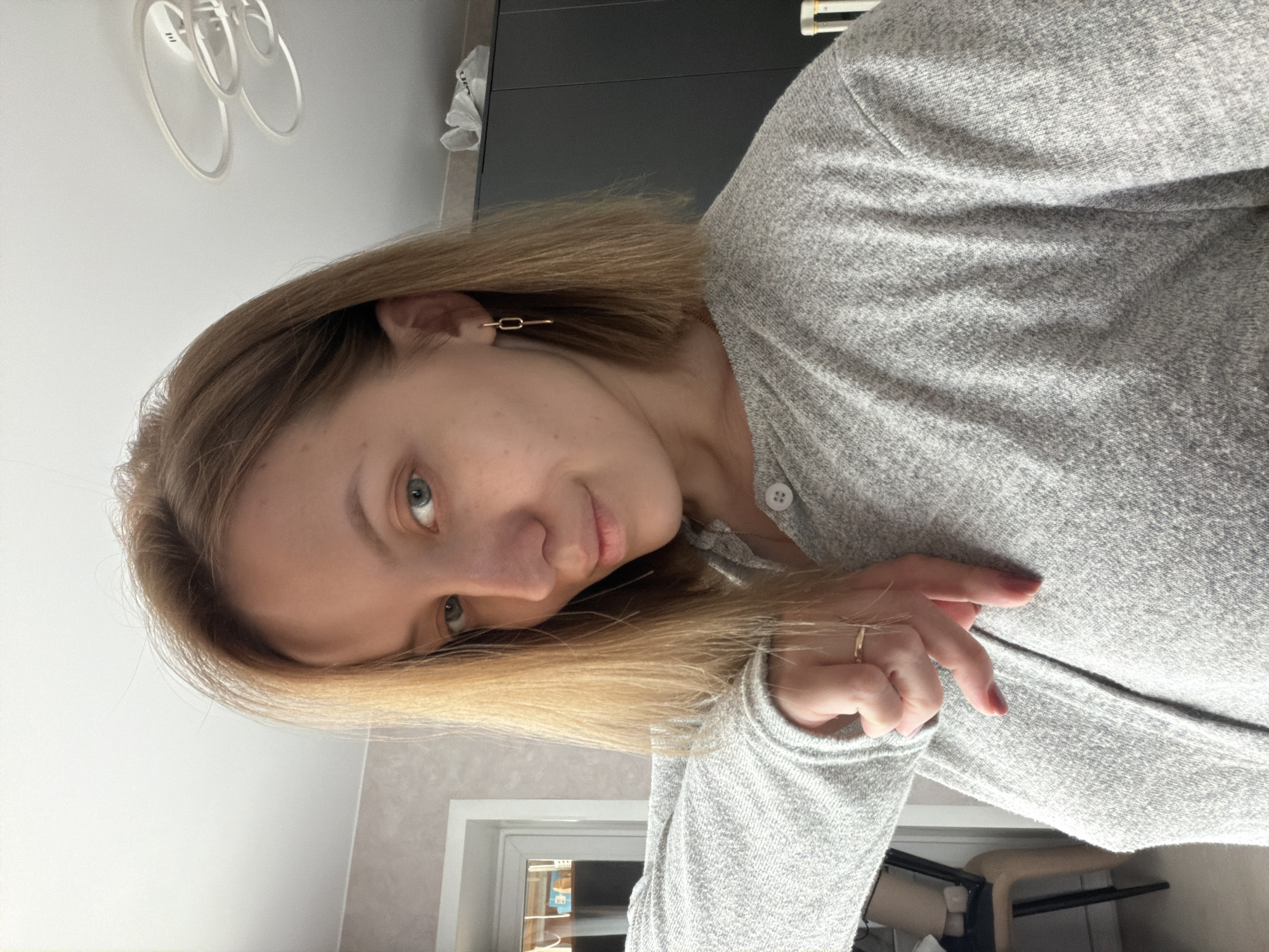 A woman with blonde hair and light eyes is seen indoors. She is wearing a grey long-sleeved top and a gold earring. She is also wearing a ring. The background is blurry but suggests a room interior with a window and some furniture. The location is not discernible as being in Reshetnikovo, Russia.
