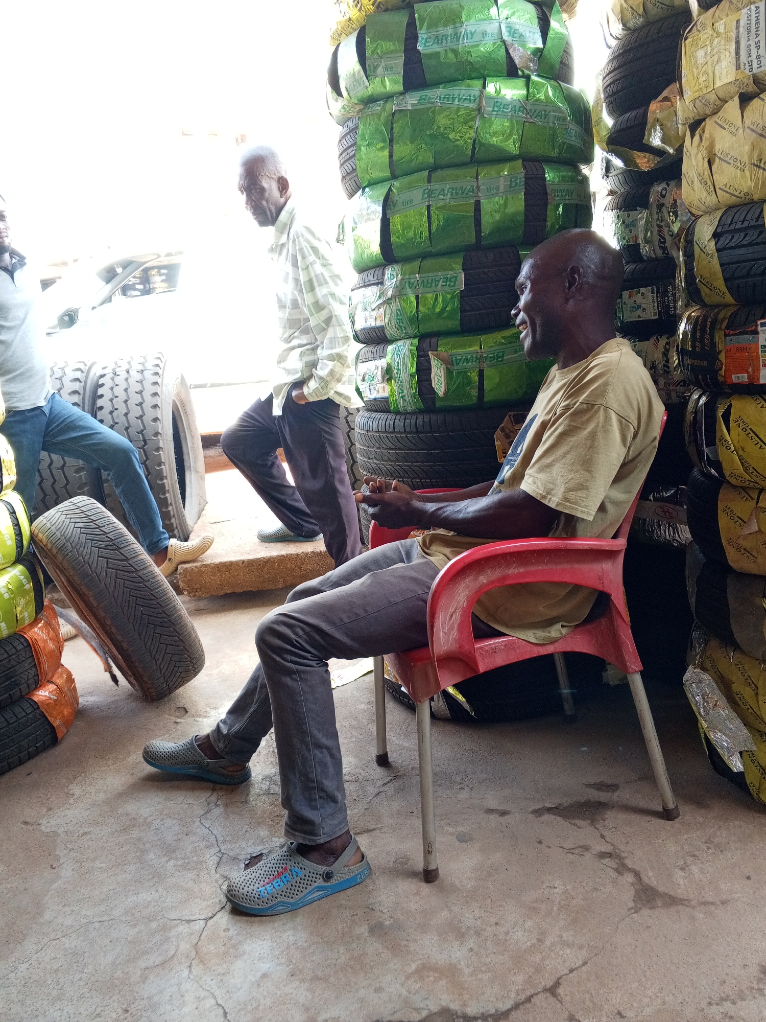 The image shows a tire shop in Maiburiji, Nigeria. A Black man wearing a tan t-shirt and gray pants is seated in a red plastic chair, looking to his left and smiling. Stacks of tires wrapped in green and yellow packaging rise behind him, some labeled "BEARWAY tire" and "AUSTONE TIRES" along with their respective tire sizes and specifications such as 195/65R15. To the left, two men stand near a stack of large tires. The man closest to the camera wears a light-colored shirt and dark trousers. The scene takes place on a concrete floor with visible cracks. The environment suggests a small business setting.