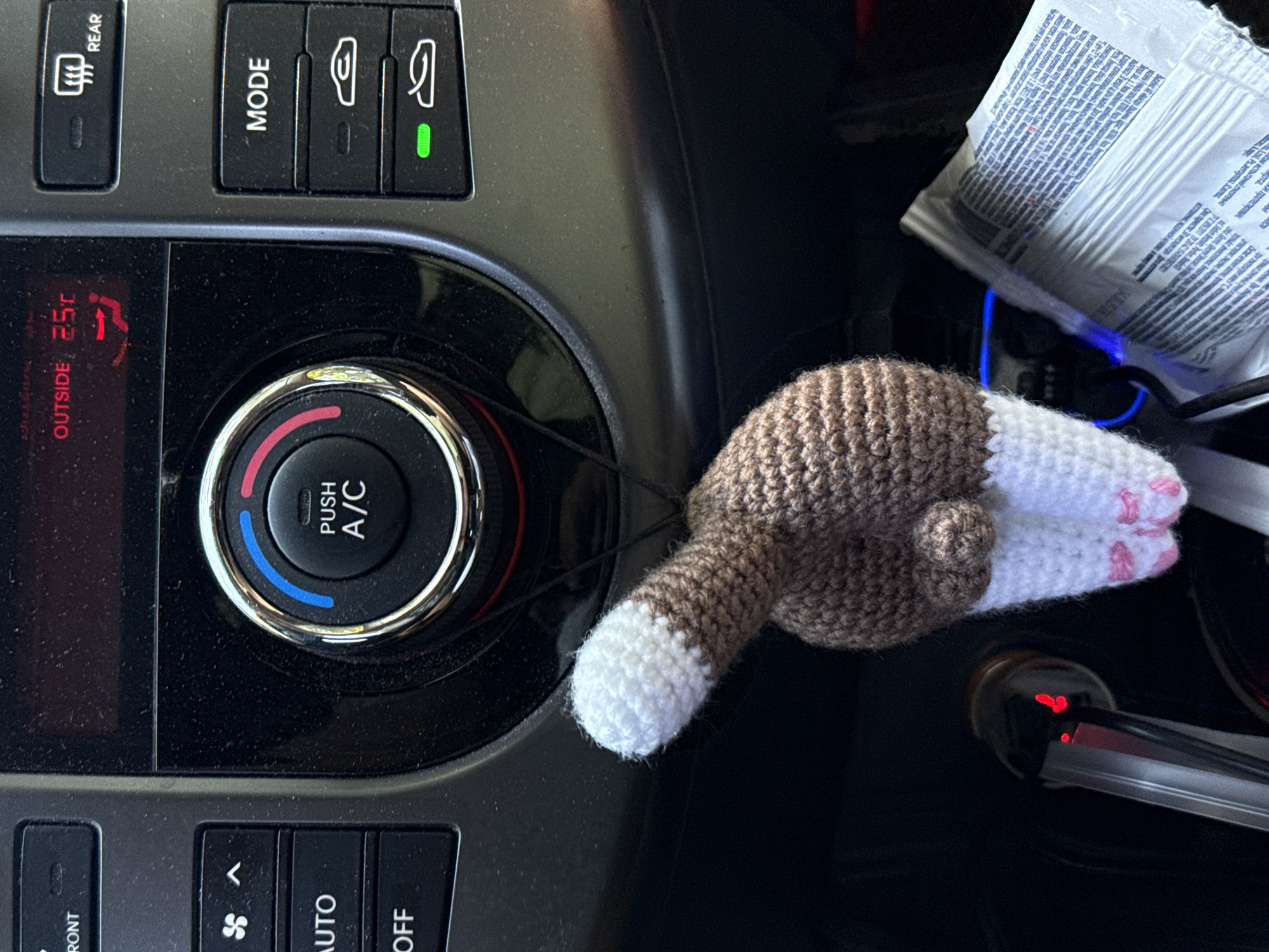 A crocheted cat tail ornament is hanging from a car's climate control knob. The car's dashboard displays "OUTSIDE 25C" with an air conditioning symbol. Buttons for "FRONT," "AUTO," and "OFF" are visible on the left, while "REAR" and "MODE" buttons are on the right. A green light is illuminated on one of the mode buttons. A car charger is plugged into the dashboard's power outlet. The background is dark, suggesting the photo was taken inside a car.